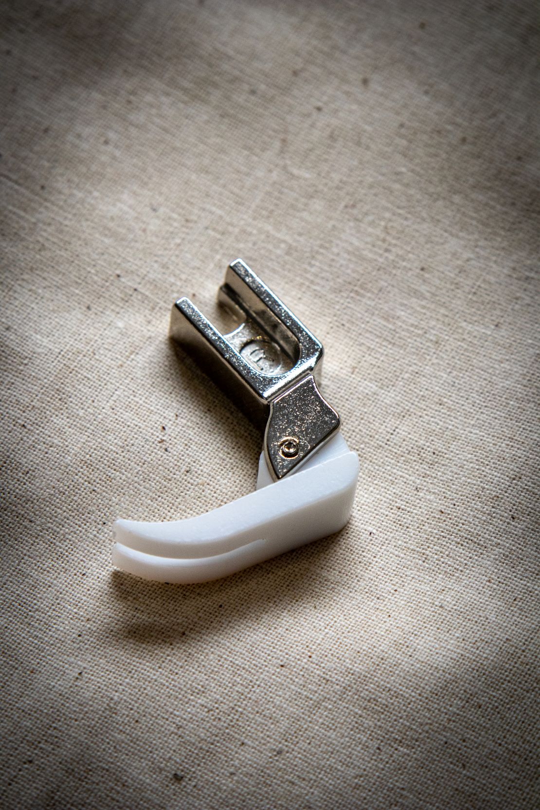 Side view of the Teflon presser foot from the RMIT Store, showing a clear plastic upper section and white non-stick base, positioned upright on a beige fabric background.