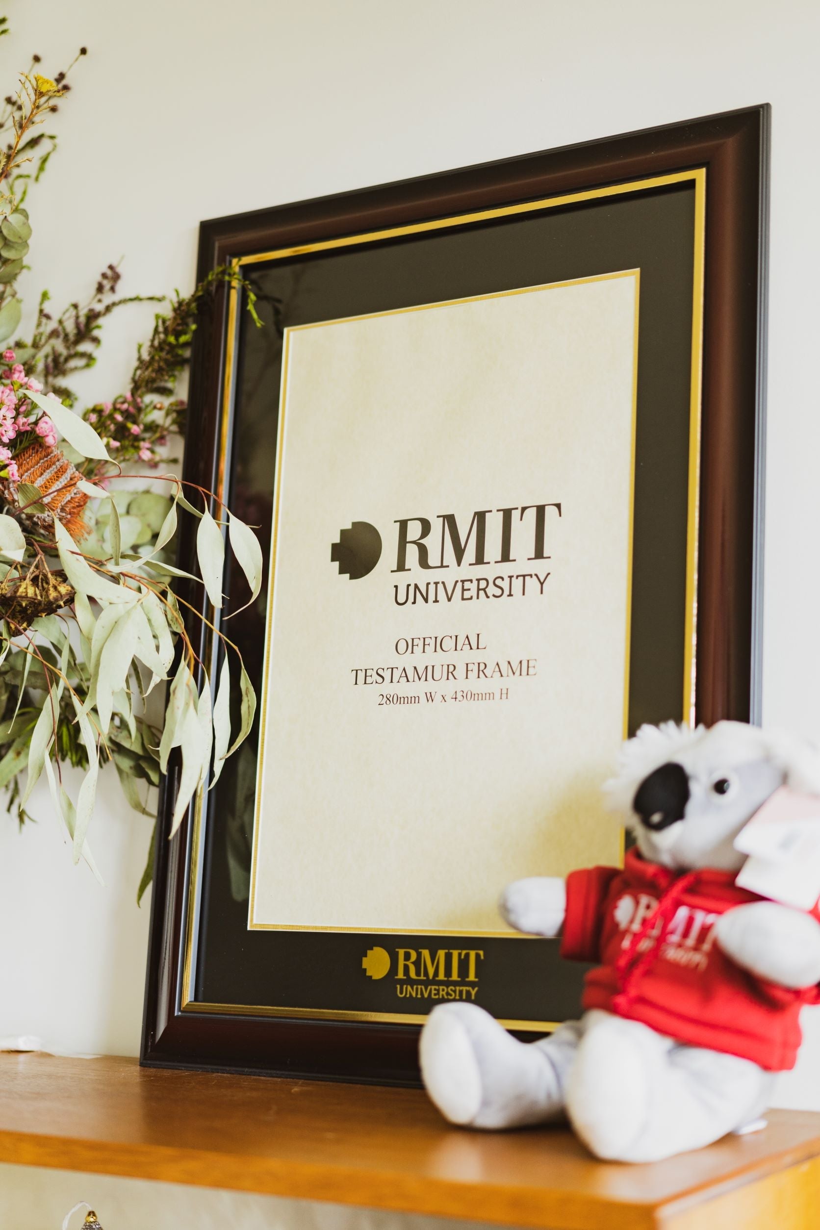 Testurmar Frame with a chestnut wood finish displayed upright on a light wooden tabletop, angled slightly to the left. The frame features a charcoal matte backing with fine gold accent lines and bold gold RMIT University branding clearly printed at the bottom of the backing. A small bunch of colourful flowers appears on the left, and a soft plushie is placed on the right. Product shown in a styled tabletop setting for the RMIT Store.