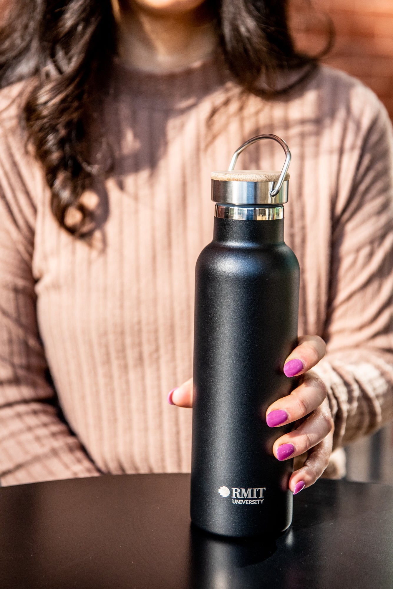 Model holding the black RMIT Store 600ml thermo bottle with the lid on and stainless steel handle raised. The bottle is upright and resting on a black tabletop, with the model’s hands positioned gently around it.