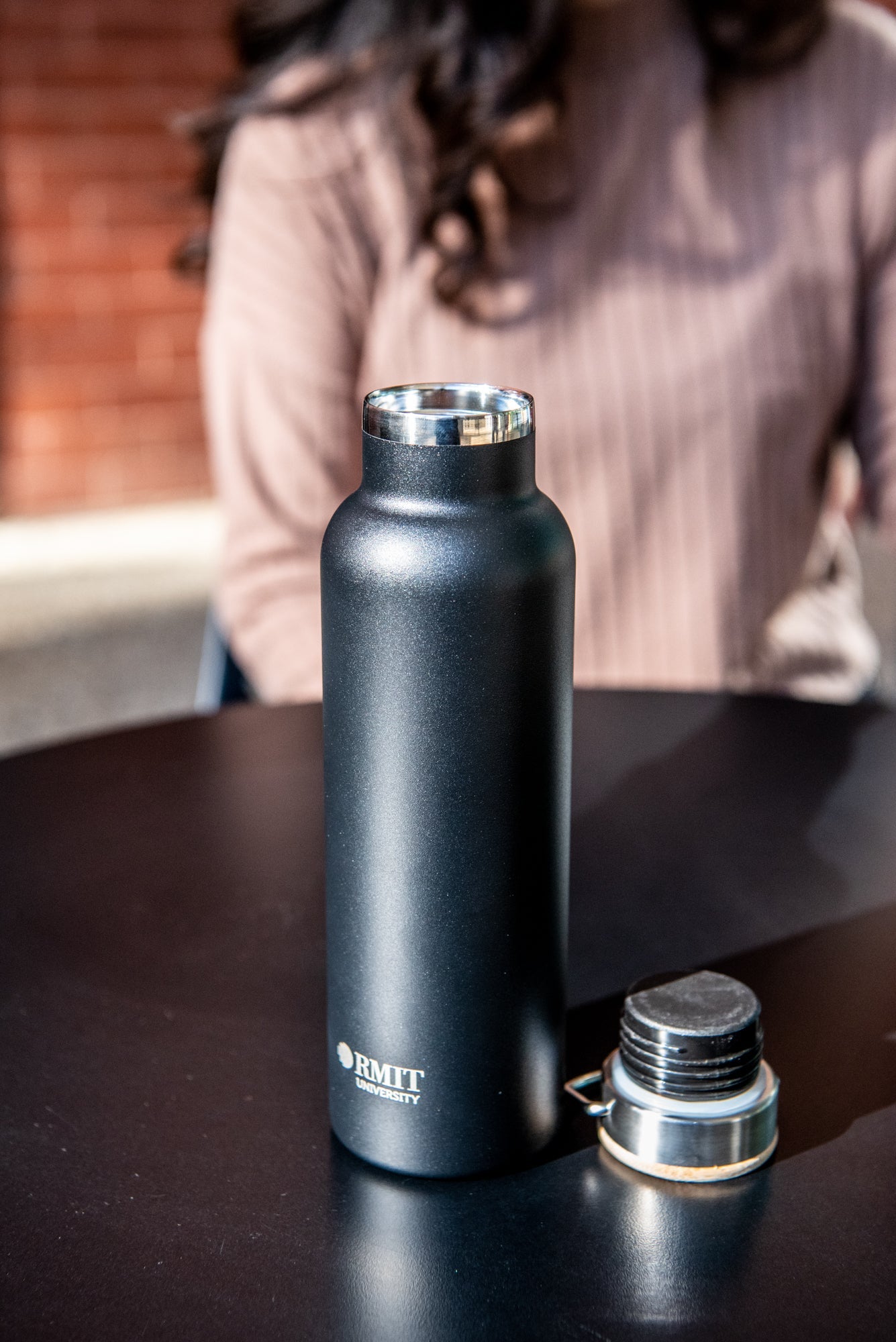Black RMIT Store 600ml thermo bottle placed upright on a black tabletop, with the natural wood lid positioned separately to the right. The 