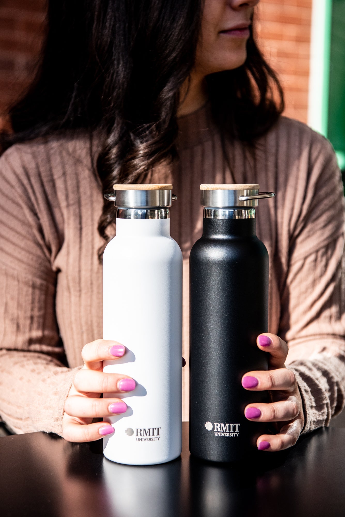 Close-up of a model’s hands resting on a black tabletop, holding two RMIT Store 600ml thermo bottles — one black and one white — each with a natural wood lid. The “RMIT University” logo in light gold is printed horizontally near the base of both bottles. 