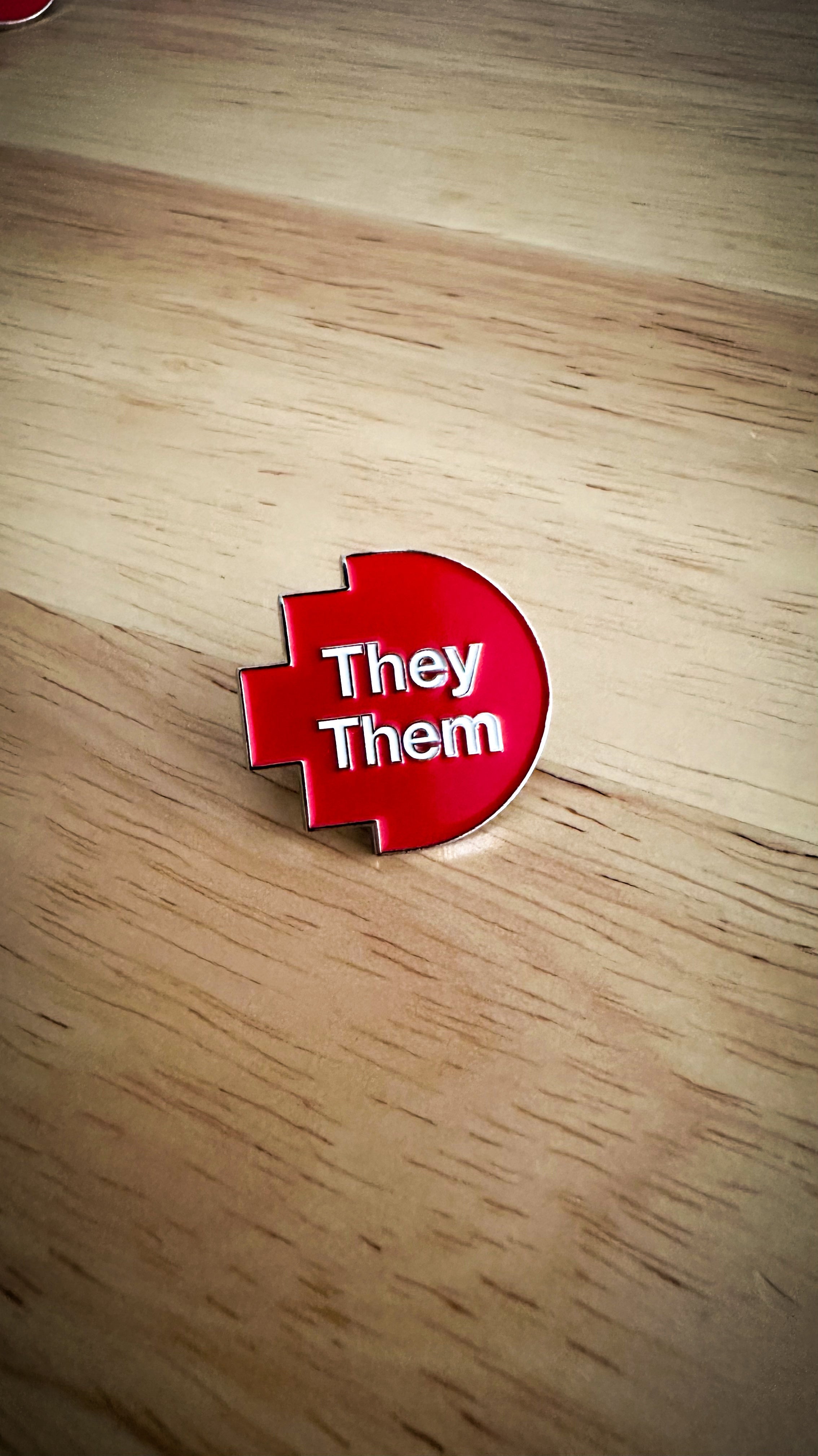 Close-up of a they/them pronoun pin from the RMIT Store, featuring white printed text “They/Them” on a red pixelated background. The pin is made of metal with a smooth finish and is displayed on a wooden surface background.