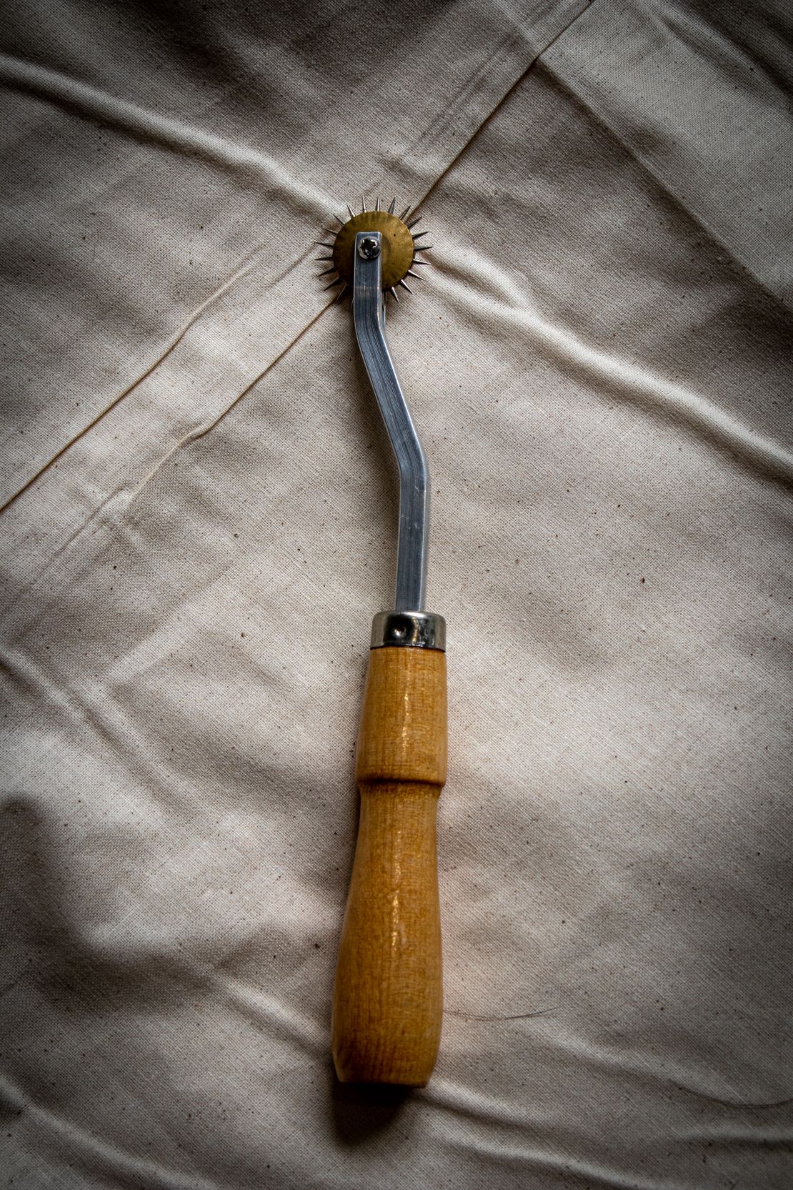 Full view of a tracing wheel with a wooden handle and metallic wheel, placed on a light grey surface with visible grain texture. Available from the RMIT Store.