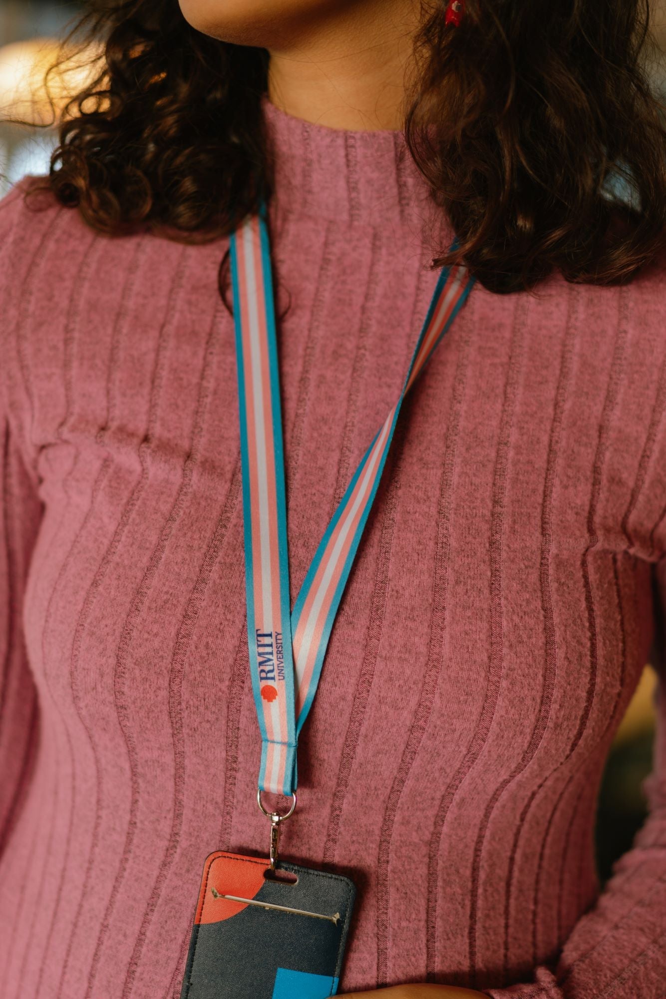 Close-up of a model wearing a Transgender Pride lanyard from the RMIT Store, featuring horizontal stripes in light blue, pink, and white with a subtle engraved RMIT logo near the silver metal clip. The lanyard is attached to a card holder. The model wears a pink top.