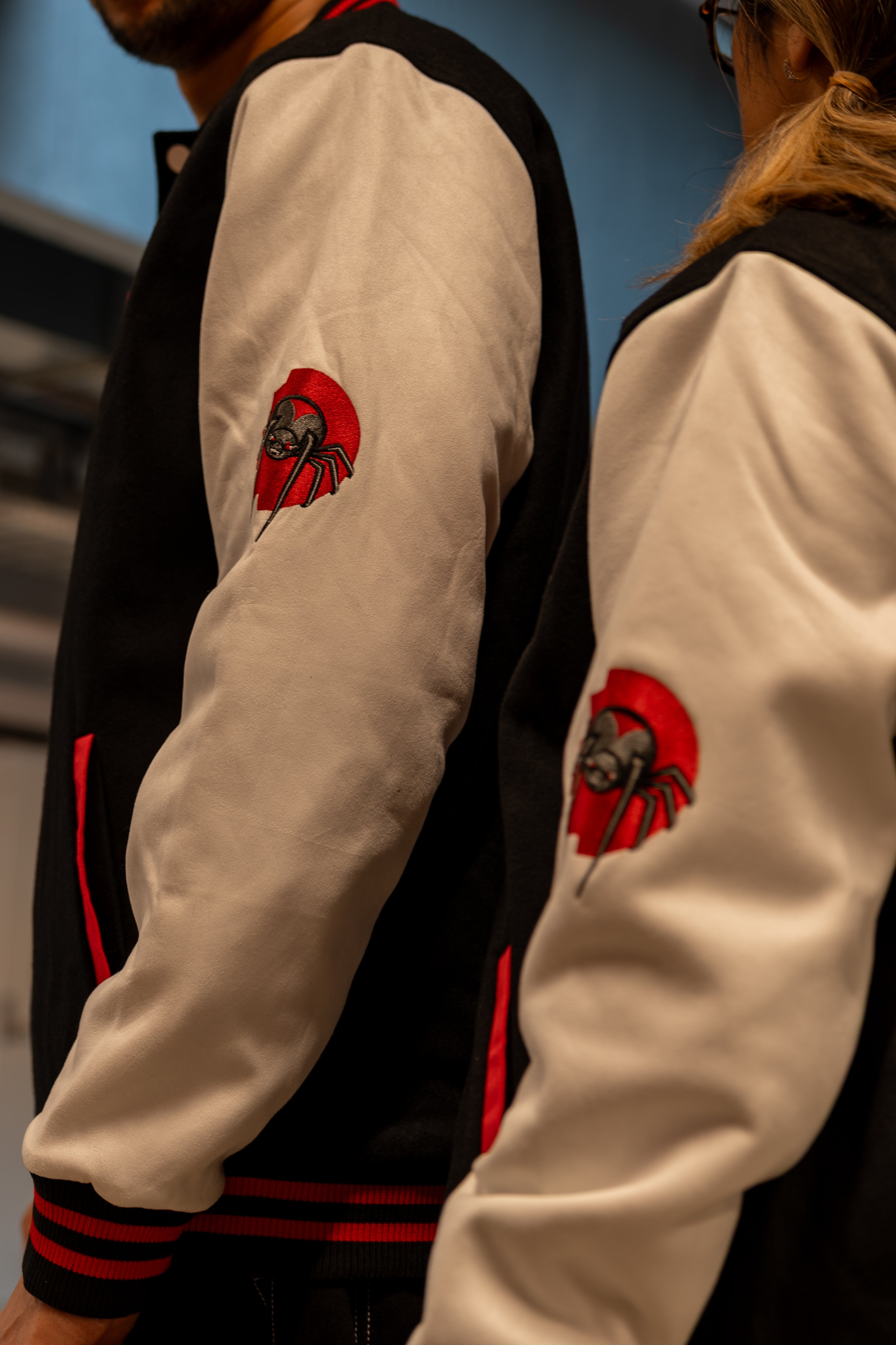Close-up side view of two people wearing the black and white RMIT varsity jacket, showing the detailed embroidered RMIT spider patch on the sleeve. The stitching and colour contrast highlight the jacket’s premium construction and signature design elements available at the RMIT Store.
