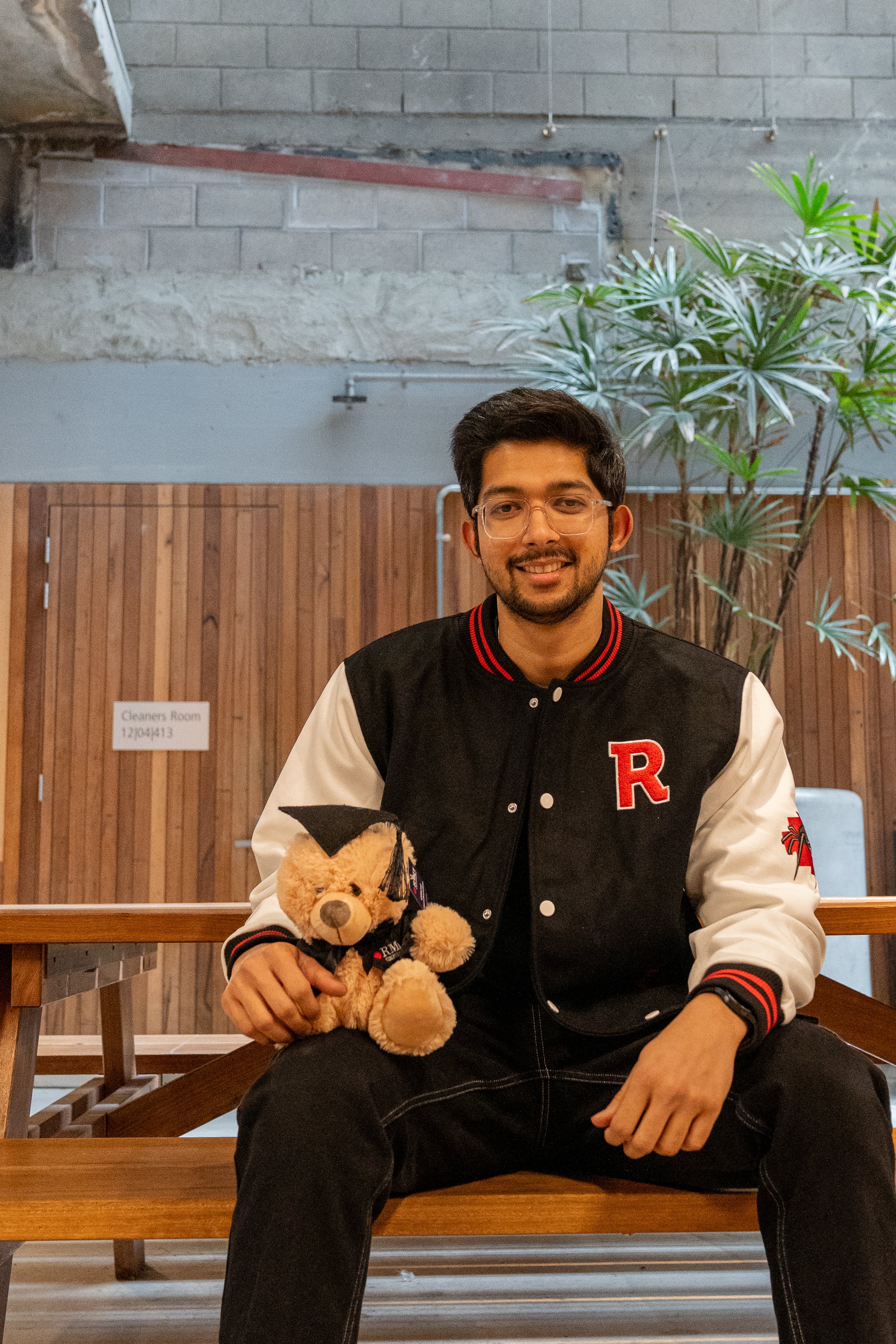 A male model wearing the black and white RMIT varsity jacket sits on a wooden bench indoors, holding the RMIT graduation teddy bear on his lap. The jacket features a bold red “R” on the chest and red-and-black striped trim, with the RMIT spider embroidery on the sleeve. Shot in a modern campus space with concrete and timber details, this product image showcases the varsity jacket available at the RMIT Store.
