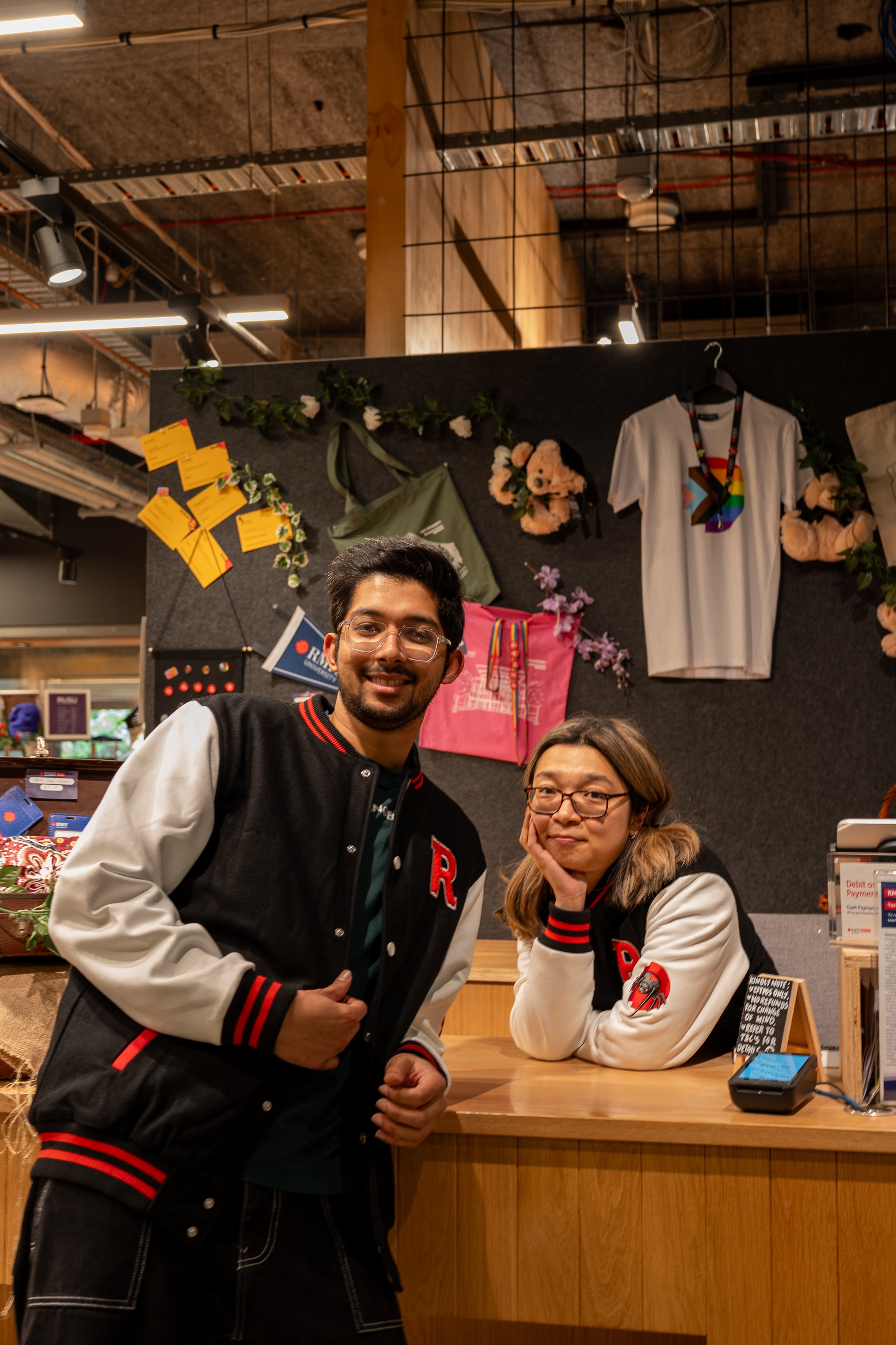 Two models wearing matching black and white RMIT varsity jackets pose inside the RMIT Store, standing at the counter with branded merchandise displayed behind them. The jackets feature the red “R” chest logo, striped collar and cuffs, and white sleeves with the embroidered RMIT spider patch. This lifestyle shot highlights the jacket’s casual campus styling and branding, available at the RMIT Store.