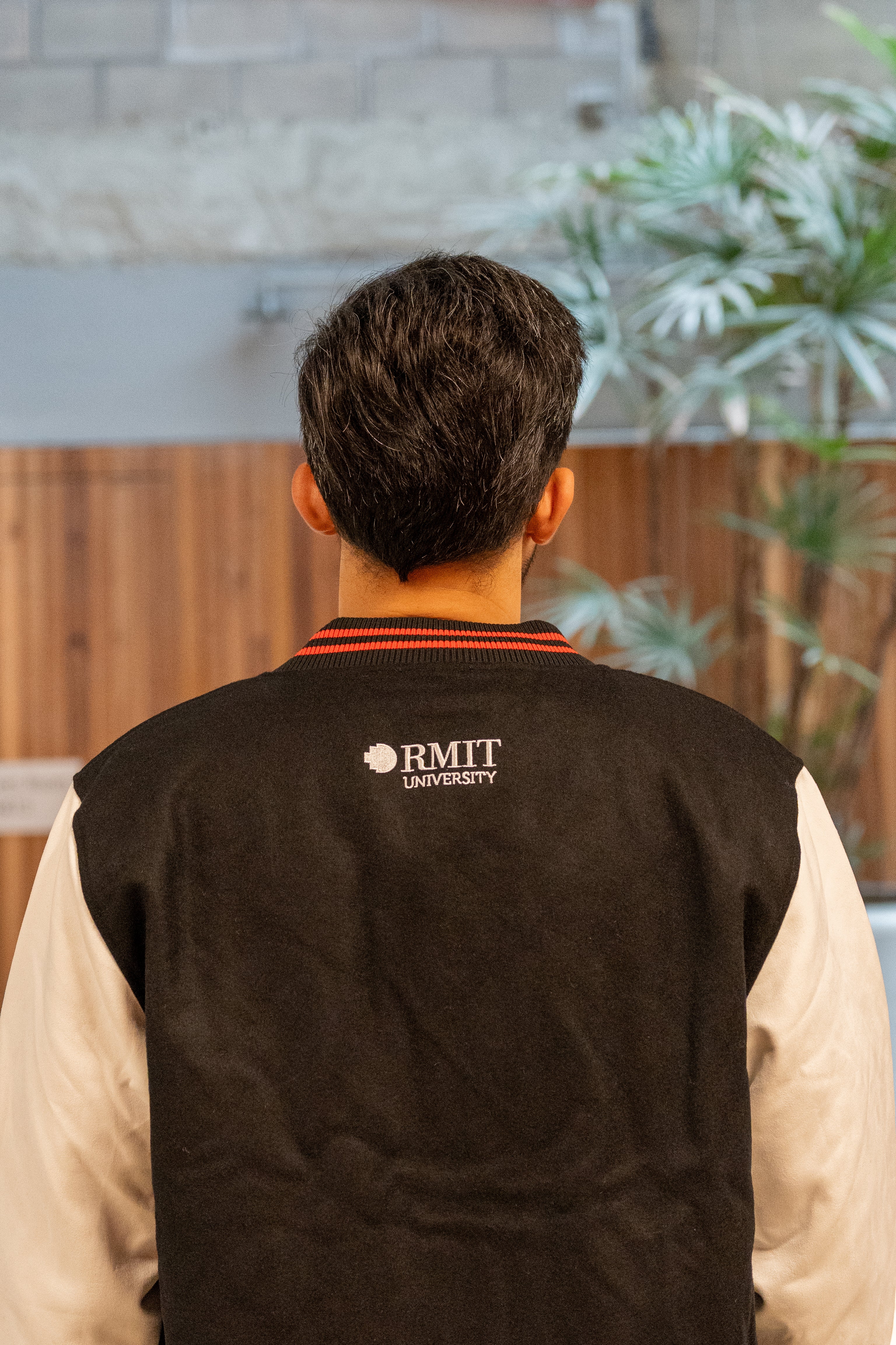 Rear view of a model wearing the black and white RMIT varsity jacket, showcasing the embroidered white RMIT University logo across the upper back. The image highlights the jacket’s clean black wool body and contrast sleeves, photographed in a modern campus setting. Available at the RMIT Store.