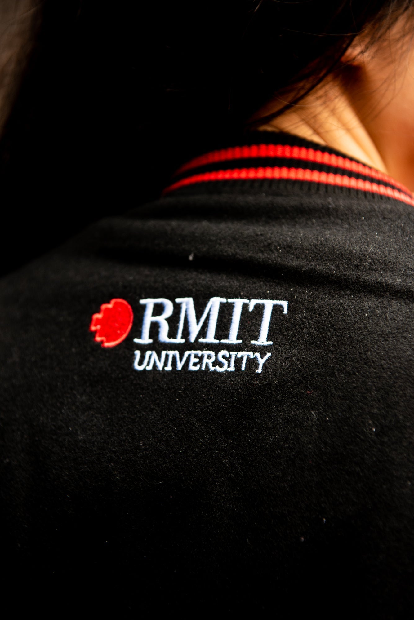 Zoomed-in view of the back collar of the black and red RMIT varsity jacket worn by a model, showing the white RMIT University logo centred behind the neck on the collar, available at the RMIT Store.