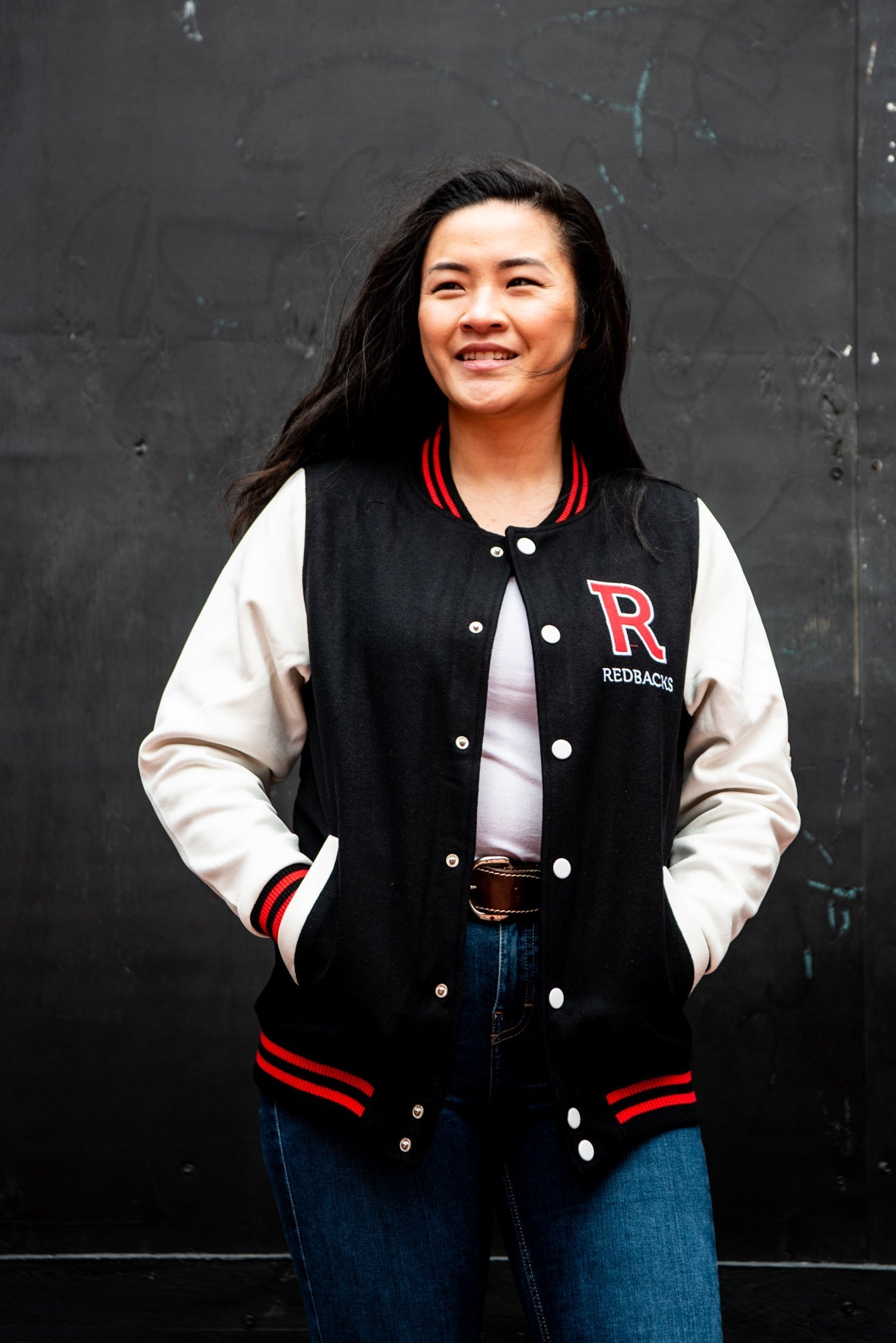 Front close-up view of female model wearing the RMIT varsity jacket, looking to her right. The black and red body and white sleeves of the jacket are visible, along with the red 'R' patch on the left chest, available at the RMIT Store.