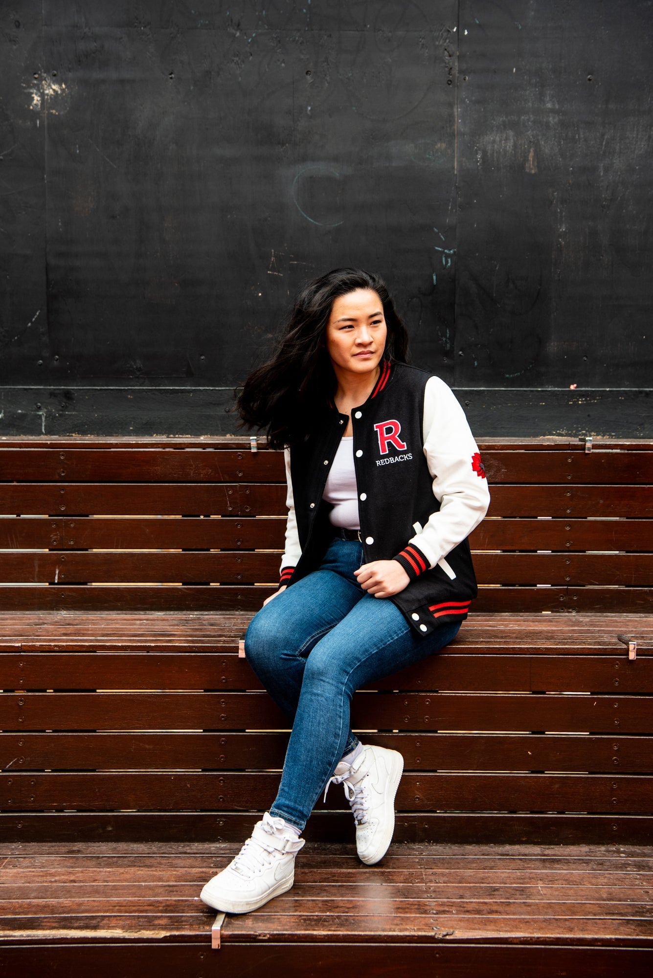 Female model sitting on a bench, looking to her right, wearing a black and red RMIT varsity jacket with white sleeves and red trim, paired with light blue denim jeans. The scene is outdoors, available at the RMIT Store.