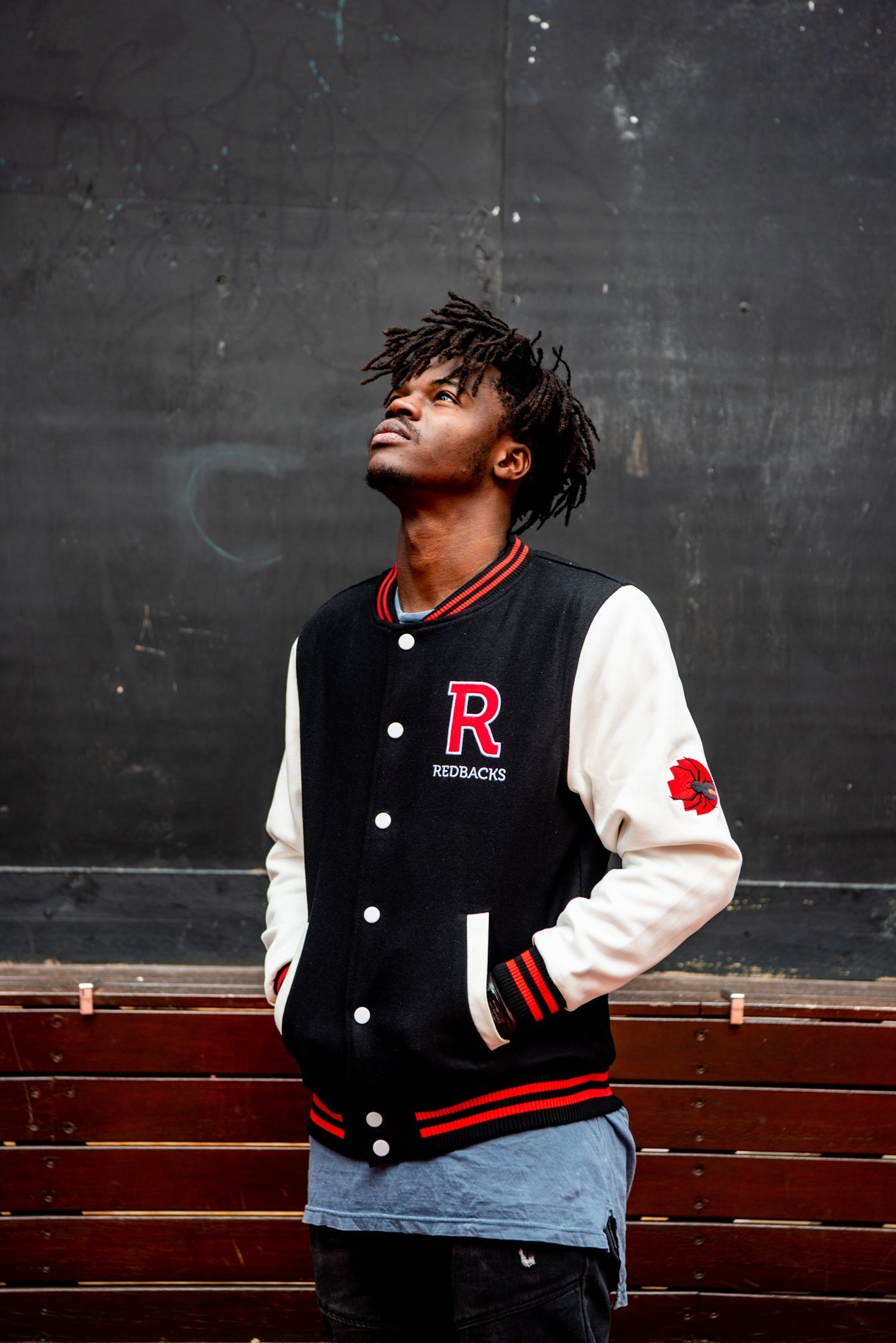 Male model wears an RMIT Store black varsity jacket with white sleeves, black collar and hem featuring red stripe detailing, and a large red chenille letter "R" patch on the left chest; white snap buttons are visible down the front as the model poses in an angled front view, looking upward at a 45-degree angle