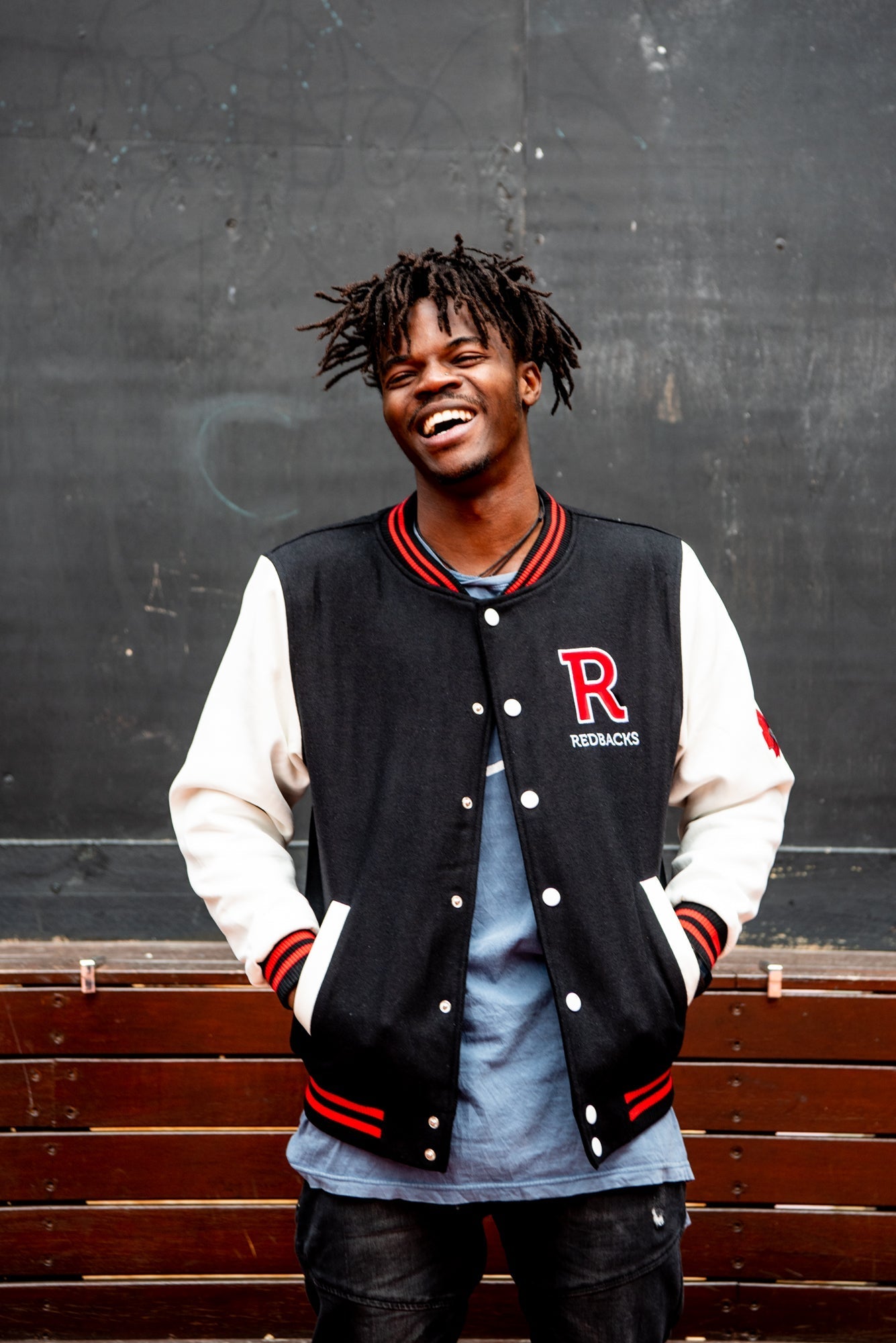 Male model wears an unbuttoned RMIT Store black varsity jacket with white sleeves, black collar and hem with red stripe accents, and a large red chenille letter "R" patch on the left chest; the model is smiling in a relaxed front-facing pose in a studio setting, revealing a blue inner shirt beneath the jacket.