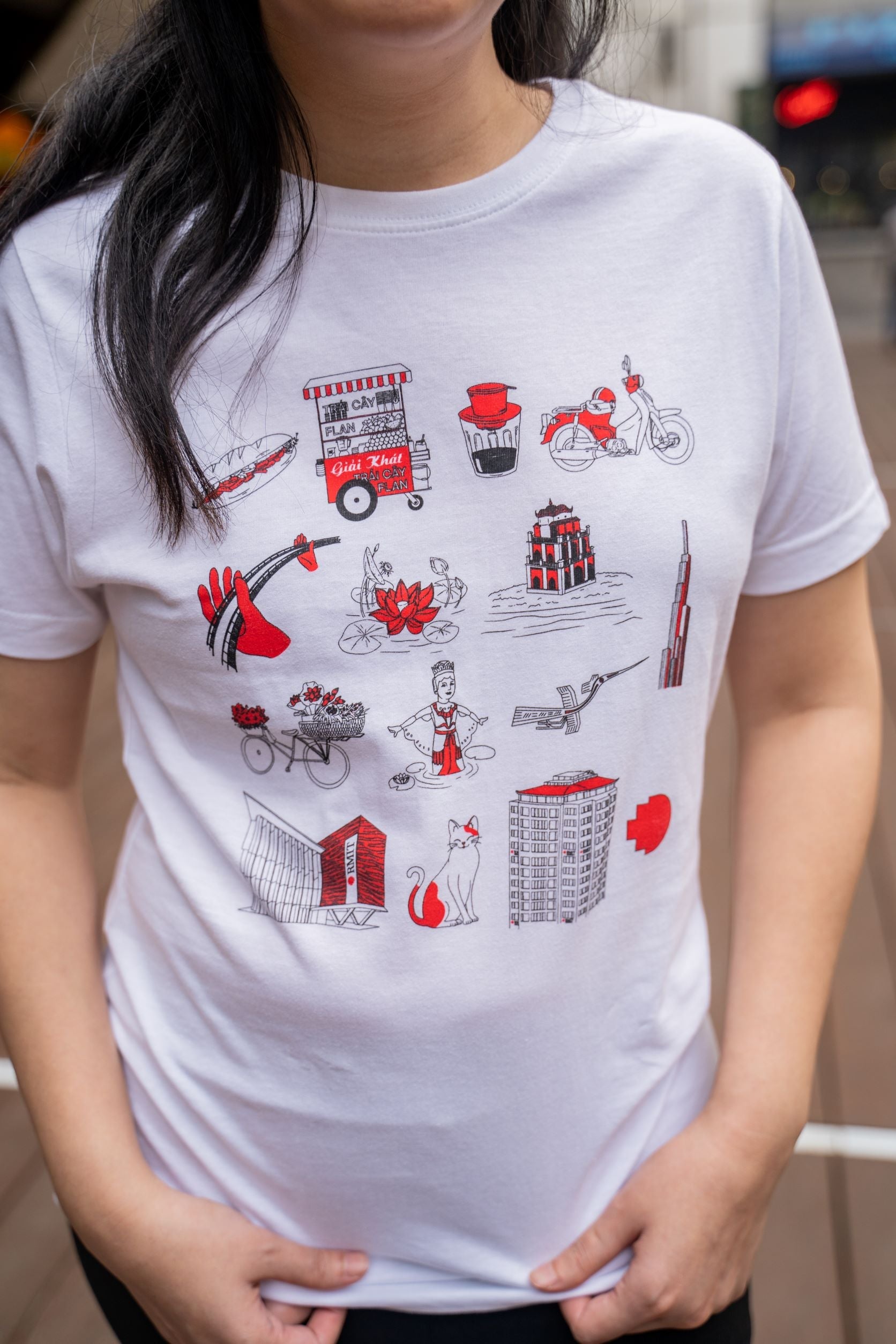 Close-up of the Vietnamese Icon Tee from RMIT Store, worn by a female model. The white shirt is adorned with red and black illustrations depicting Vietnamese cultural elements such as a food cart, traditional motifs, and notable buildings.