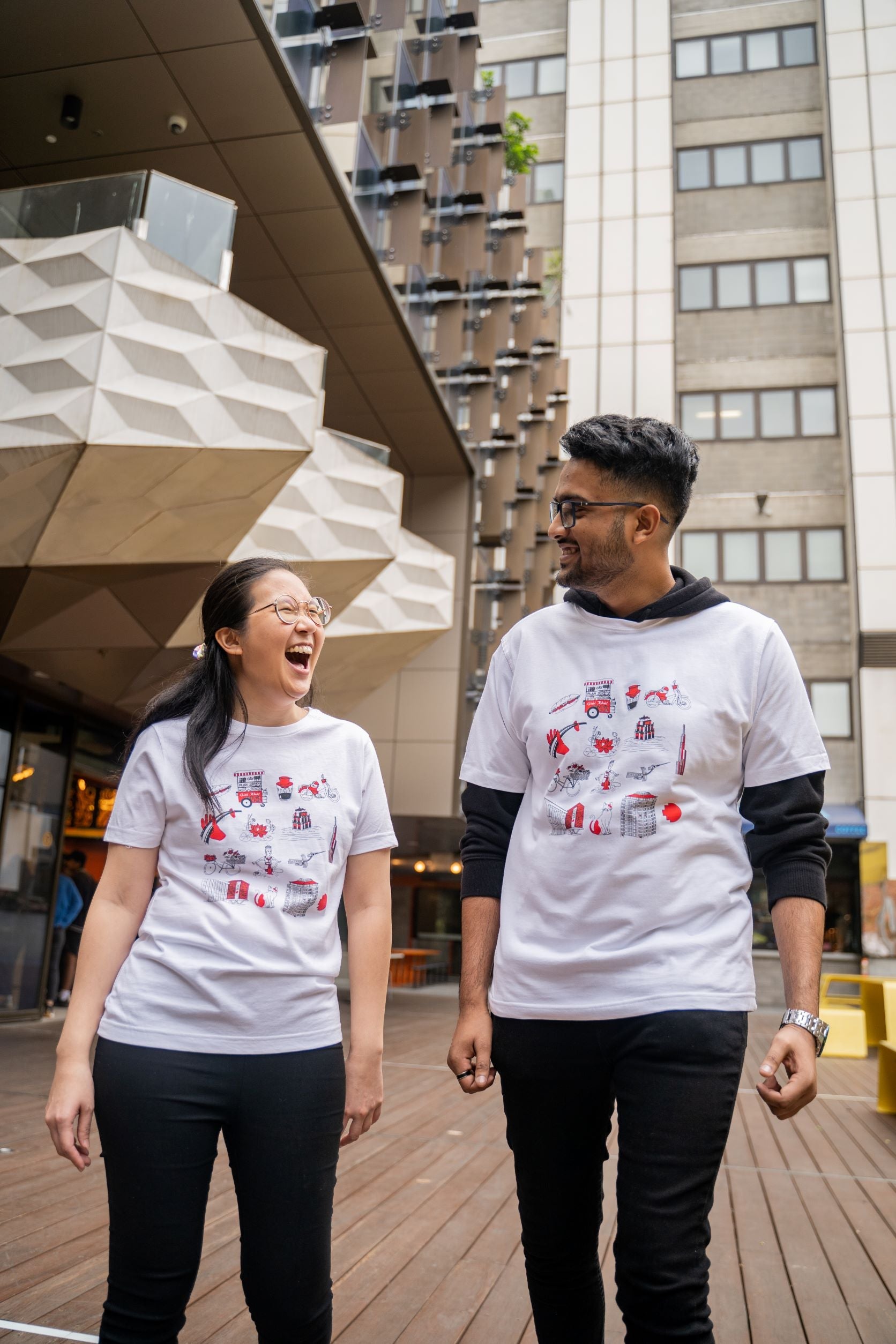 Two models walking on a wooden deck in an urban setting, wearing the Vietnamese Icon Tee from RMIT Store. The T-shirts feature red and black Vietnamese-inspired designs including cultural icons and architecture. Modern buildings with geometric features provide a striking backdrop.
