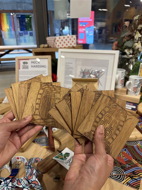 We Scar Many Trees Square Coasters Set of 9 featuring laser-engraved Indigenous artwork on dark wood square coasters, stacked and fanned on a timber display table in-store at RMIT Store, showing multiple intricate line and pattern designs with visible etched detailing and natural wood grain, with other wooden boards and cultural artwork surrounding the display.