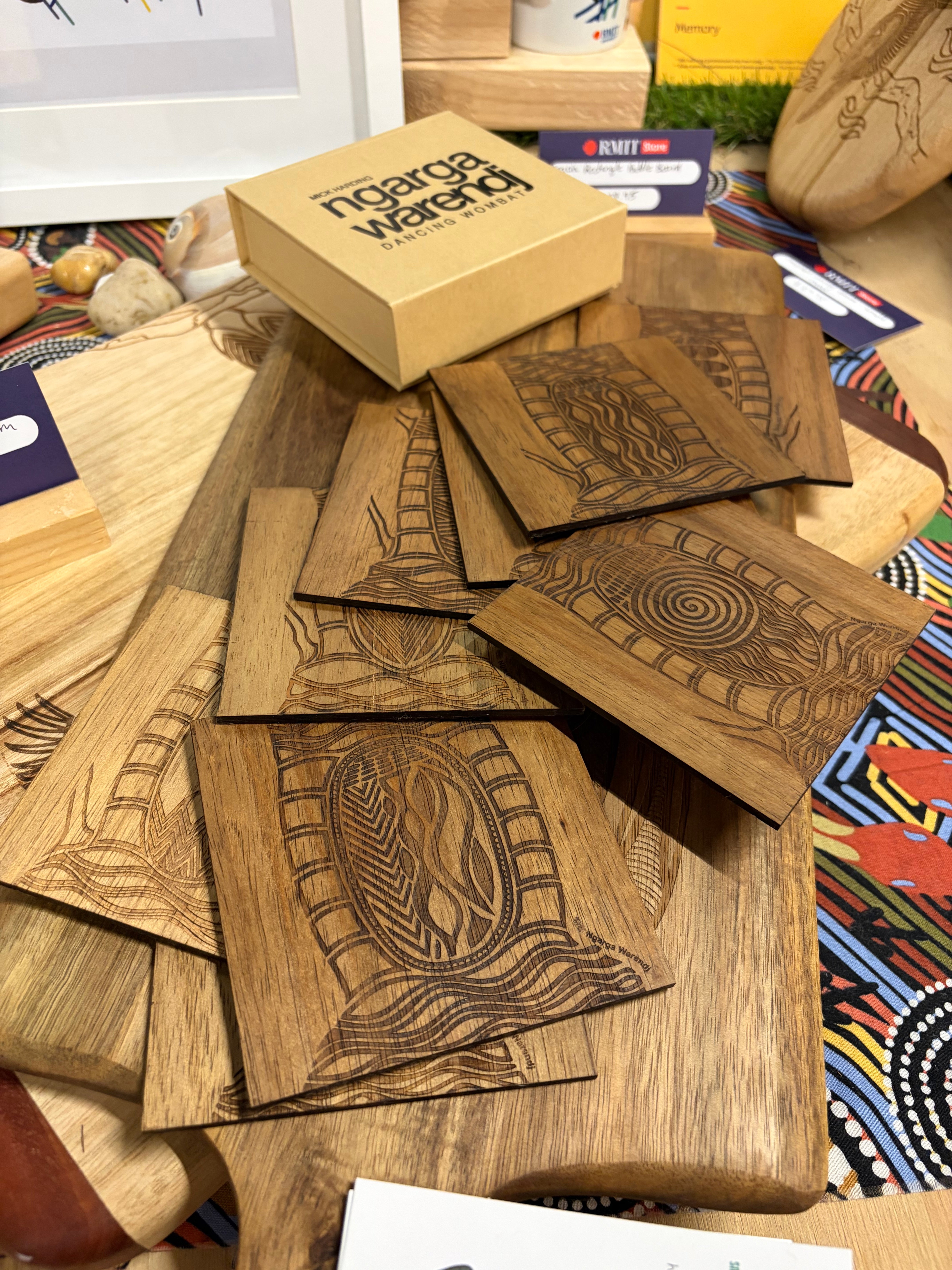 We Scar Many Trees Square Coasters – Set of 9, shown stacked and overlapping on a wooden display board in an in-store setting at RMIT Store, featuring square medium-brown timber coasters with fine laser-etched Aboriginal linework designs, including concentric circles, flowing lines, and oval central motifs, with subtle engraved “We Scar Many Trees” branding visible near the lower edge of some coasters, surrounded by wooden boards, product cards, and a kraft presentation box in the background.