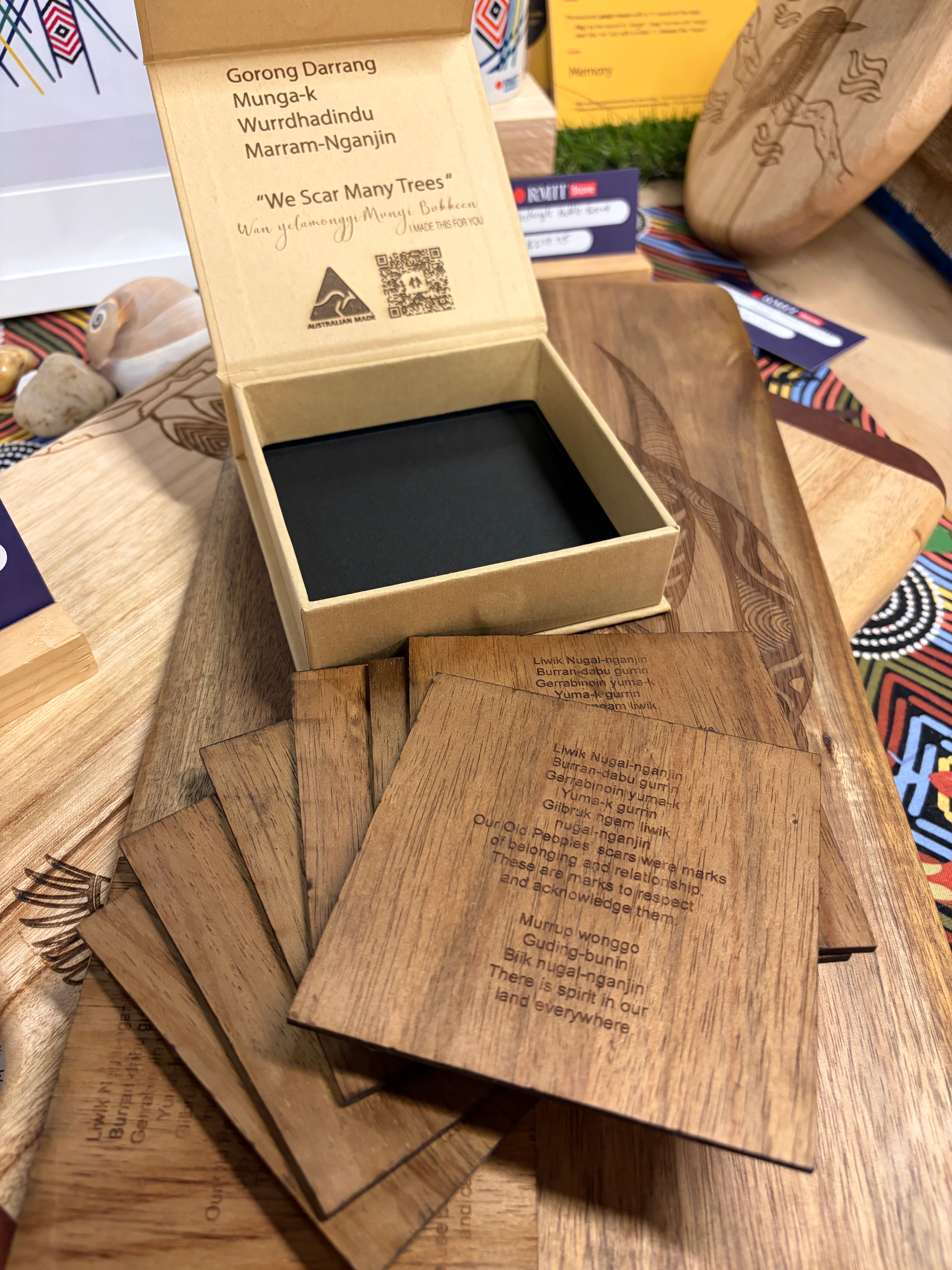 We Scar Many Trees Square Coasters – Set of 9, displayed with several coasters face down showing laser-etched text passages and Indigenous language inscriptions on smooth brown timber, placed in front of an open kraft presentation box at RMIT Store, with the box lid interior printed with the title “We Scar Many Trees,” additional Indigenous language text, a QR code, and an Australian Made kangaroo logo, set on a wooden surface among other store display items.