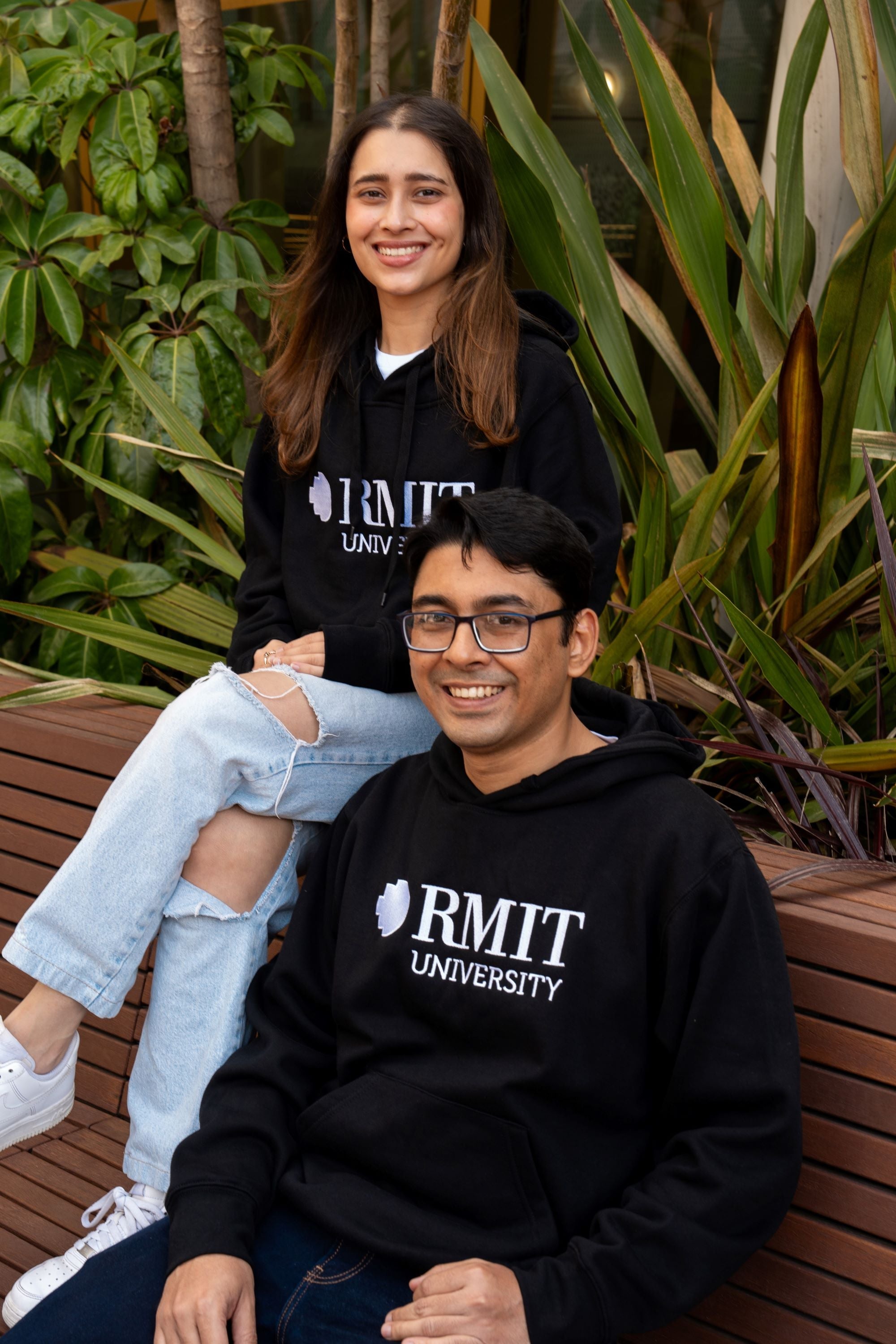 Front view of two models wearing black RMIT Welcome hoodies from the RMIT Store. The female model on the left is sitting on a higher bench, with light blue denim jeans clearly visible, while the male model on the right is seated lower. Both hoodies feature a centred white embroidered RMIT logo on the chest. 