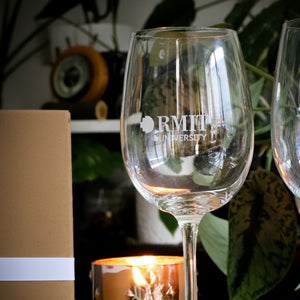  A close-up of a single wine glass etched with the RMIT University logo, showcasing the fine craftsmanship of the glass's curvature. In the background, a warm, lit candle and verdant foliage add to the serene and inviting setting. Available at the RMIT Store.