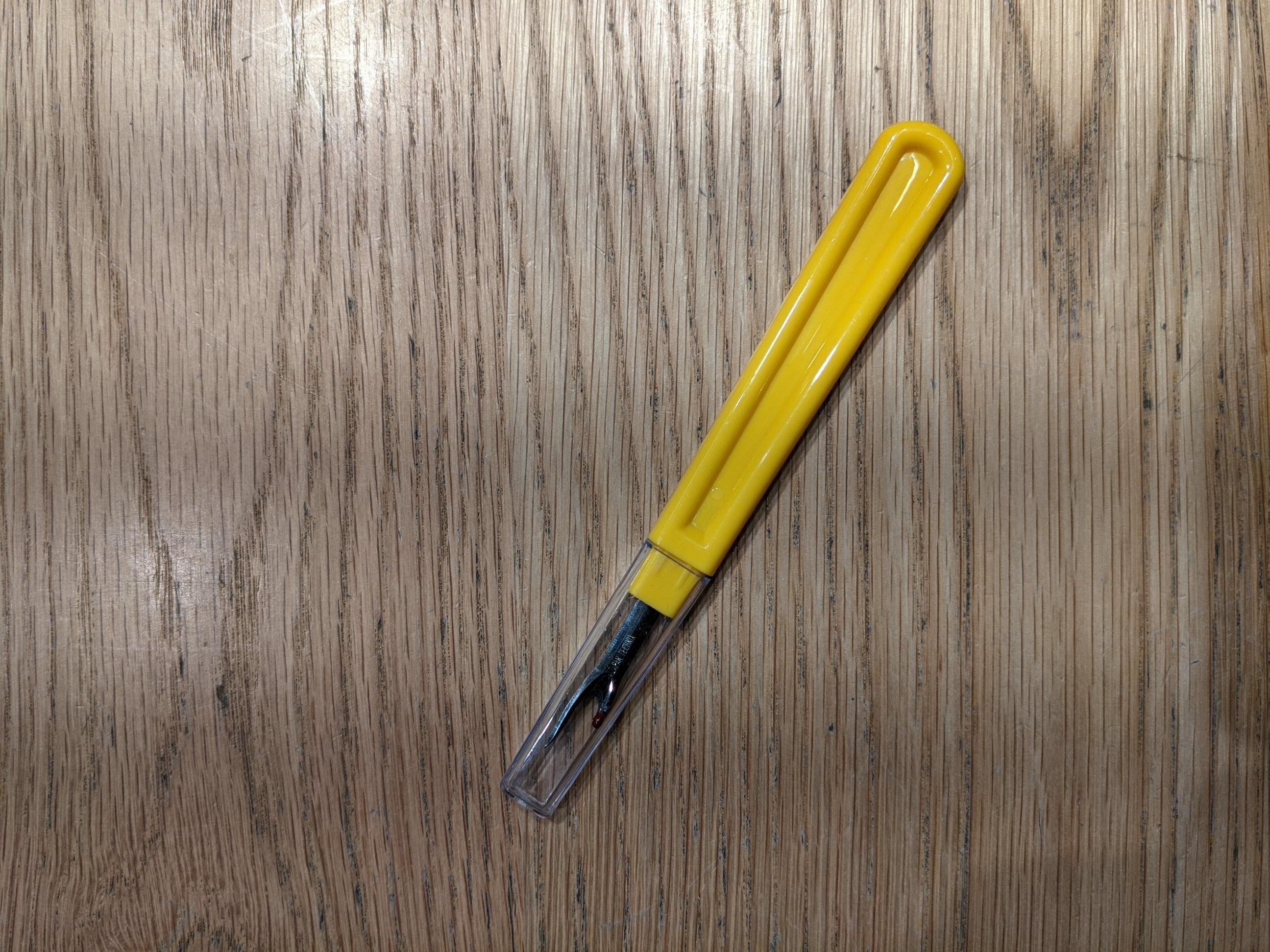 Yellow seam unpicker with a smooth curved plastic handle and transparent protective cap, displayed horizontally on a beige fabric tabletop. The exposed metal blade includes a red safety ball at the shorter tip. Sold through the RMIT Store.