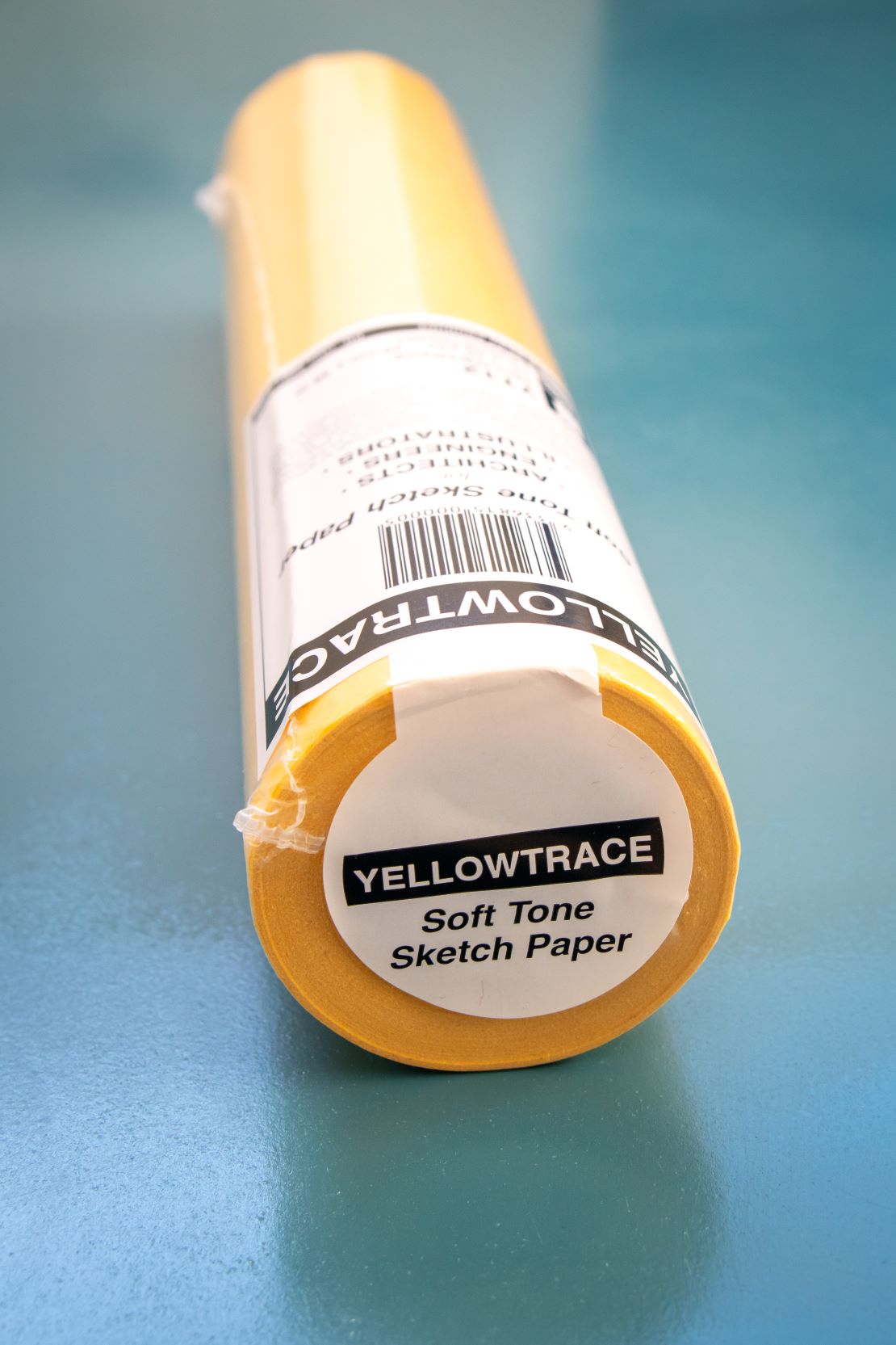 Angled view of a yellow trace paper roll with a white sticker label on the end reading “soft tone sketch paper,” showing the textured paper edge and part of the roll’s side, against a plain teal background. Available at the RMIT Store.