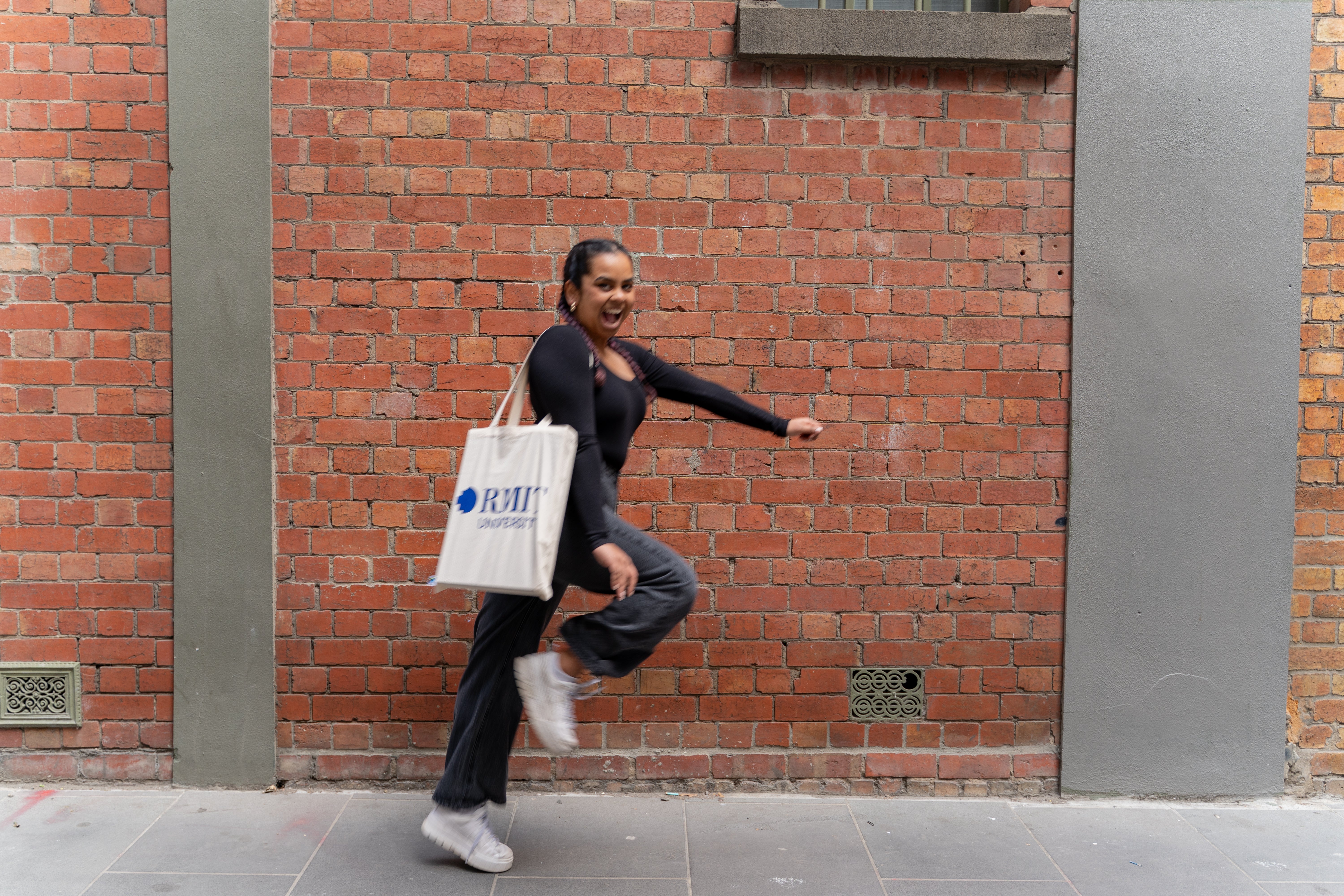 Fairtrade cotton tote bag in natural beige colour with large bold blue RMIT University logo printed on the front, carried on the shoulder by a smiling model in a playful walking pose, wearing a long-sleeve black top, dark grey trousers, and white sneakers, photographed outdoors against a full red brick wall background — available at the RMIT Store.