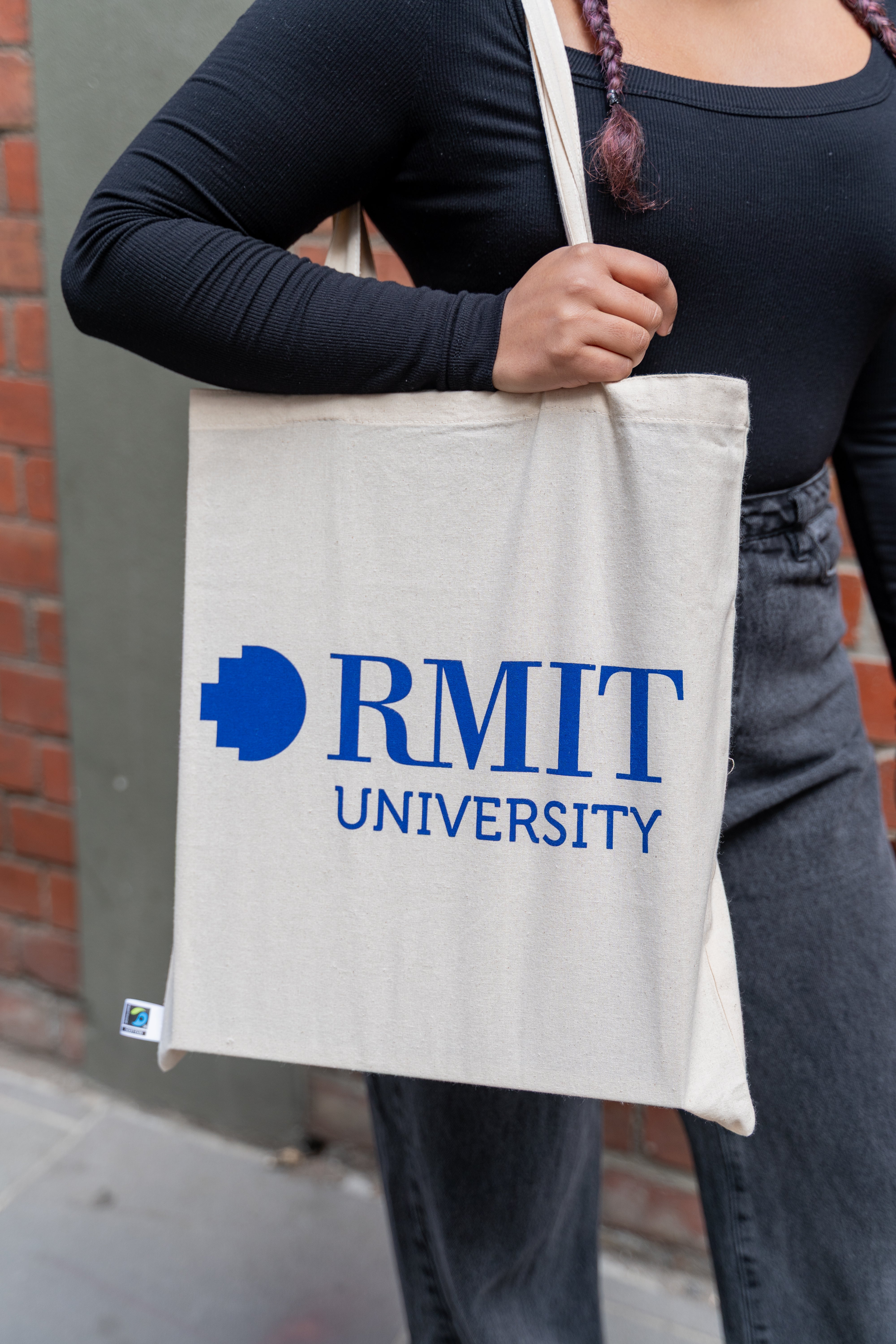 Fairtrade cotton tote bag in natural beige colour with large bold blue RMIT University logo printed on the front, carried over the shoulder by a model wearing a long-sleeve black ribbed top and dark grey trousers, with Fairtrade certification tag visible near the bottom seam, photographed outdoors against a blurred brick wall background — available at the RMIT Store.