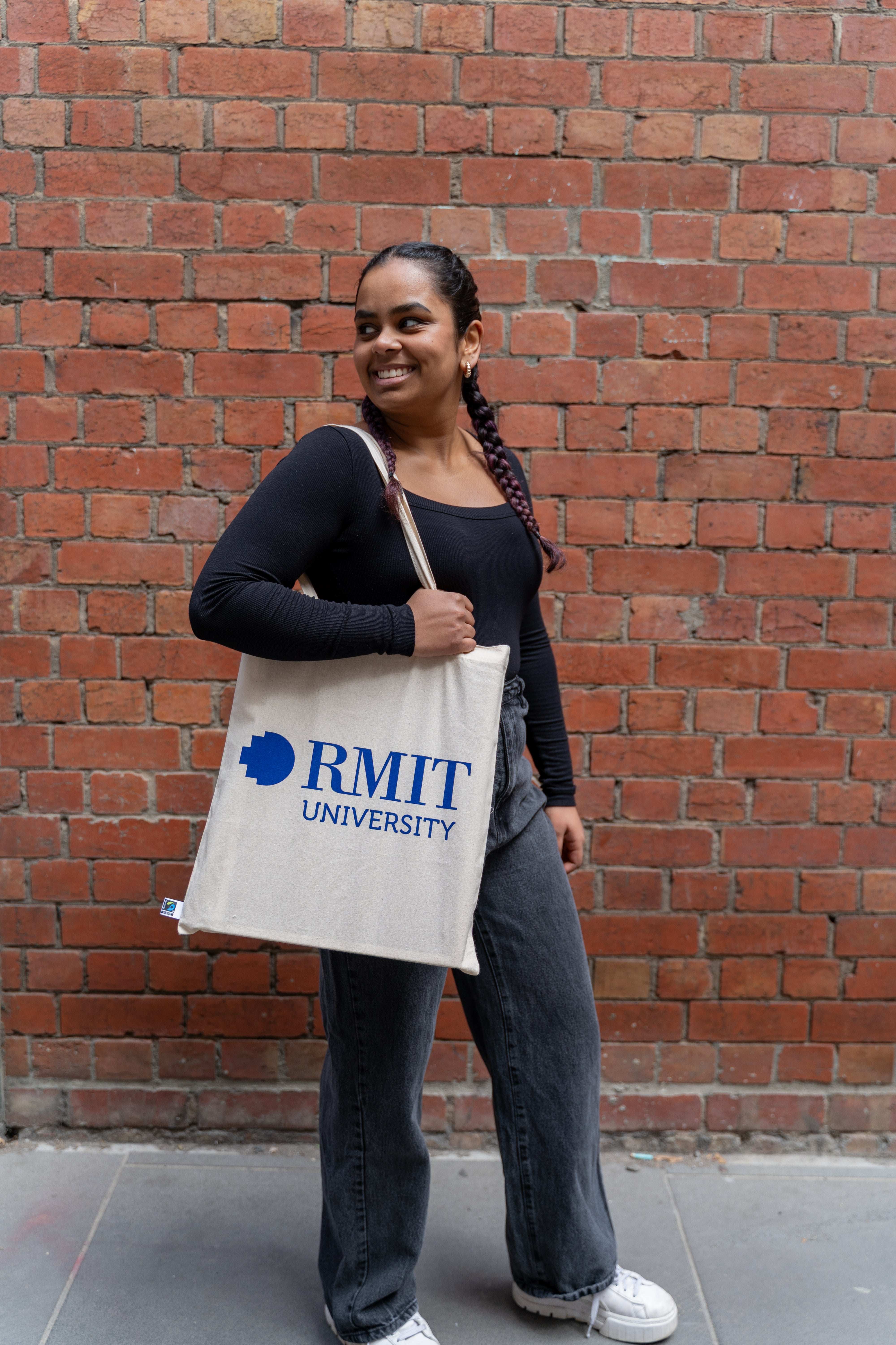 Fairtrade cotton tote bag in natural beige colour with large bold blue RMIT University logo printed on the front, carried on the shoulder by a smiling model with braided hair, wearing a long-sleeve black ribbed top and dark grey trousers, standing in front of a textured red brick wall — available at the RMIT Store.