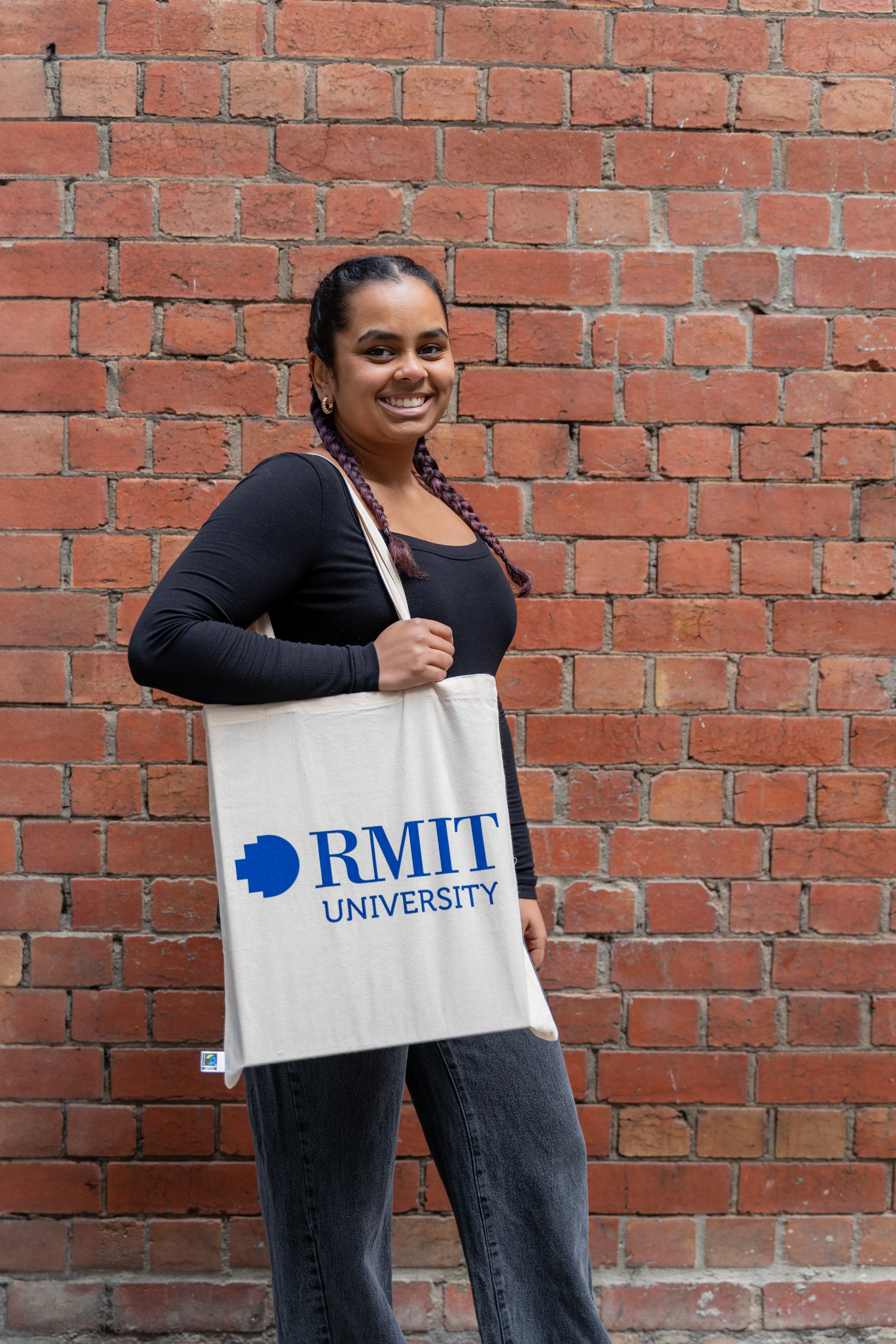 Fairtrade cotton tote bag in natural beige colour with large bold blue RMIT University logo printed on the front, carried on the shoulder by a smiling model with braided hair, wearing a long-sleeve black ribbed top and dark grey trousers, standing in front of a textured red brick wall — available at the RMIT Store.