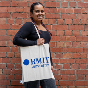 Fairtrade cotton tote bag in natural beige colour with large bold blue RMIT University logo printed on the front, carried on the shoulder by a smiling model with braided hair, wearing a long-sleeve black ribbed top and dark grey trousers, standing in front of a textured red brick wall — available at the RMIT Store.