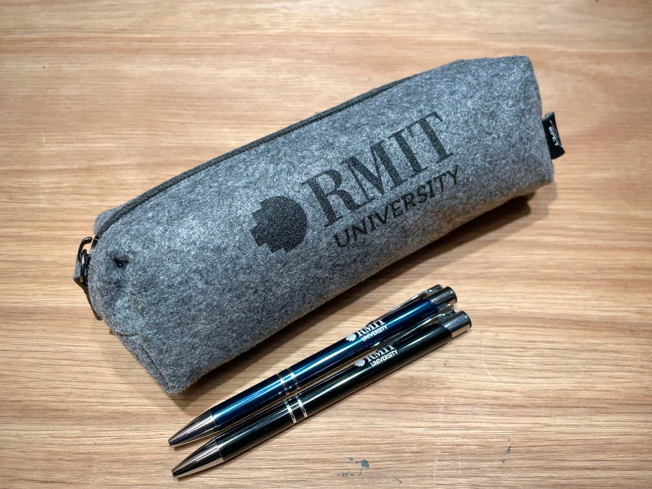 Angled front view of the RMIT Store rPET Utility Case in charcoal, displaying the black RMIT logo printed in the centre of the front panel. Two pens, one black and one navy blue, are placed in front of the case on a plain surface. The case features a textured fabric exterior with visible stitching and a closed zipper.