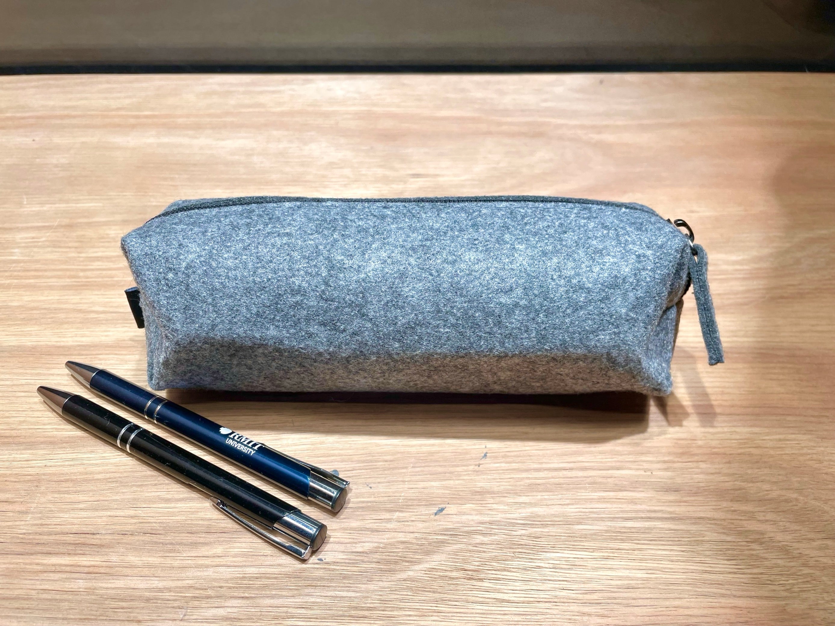 Rear view of the RMIT Store rPET Utility Case in charcoal colour. Two pens—one blue and one black—are placed next to the case on a plain surface. The case has a textured fabric exterior with visible stitching and a zipper along the edge, highlighting its durable design.