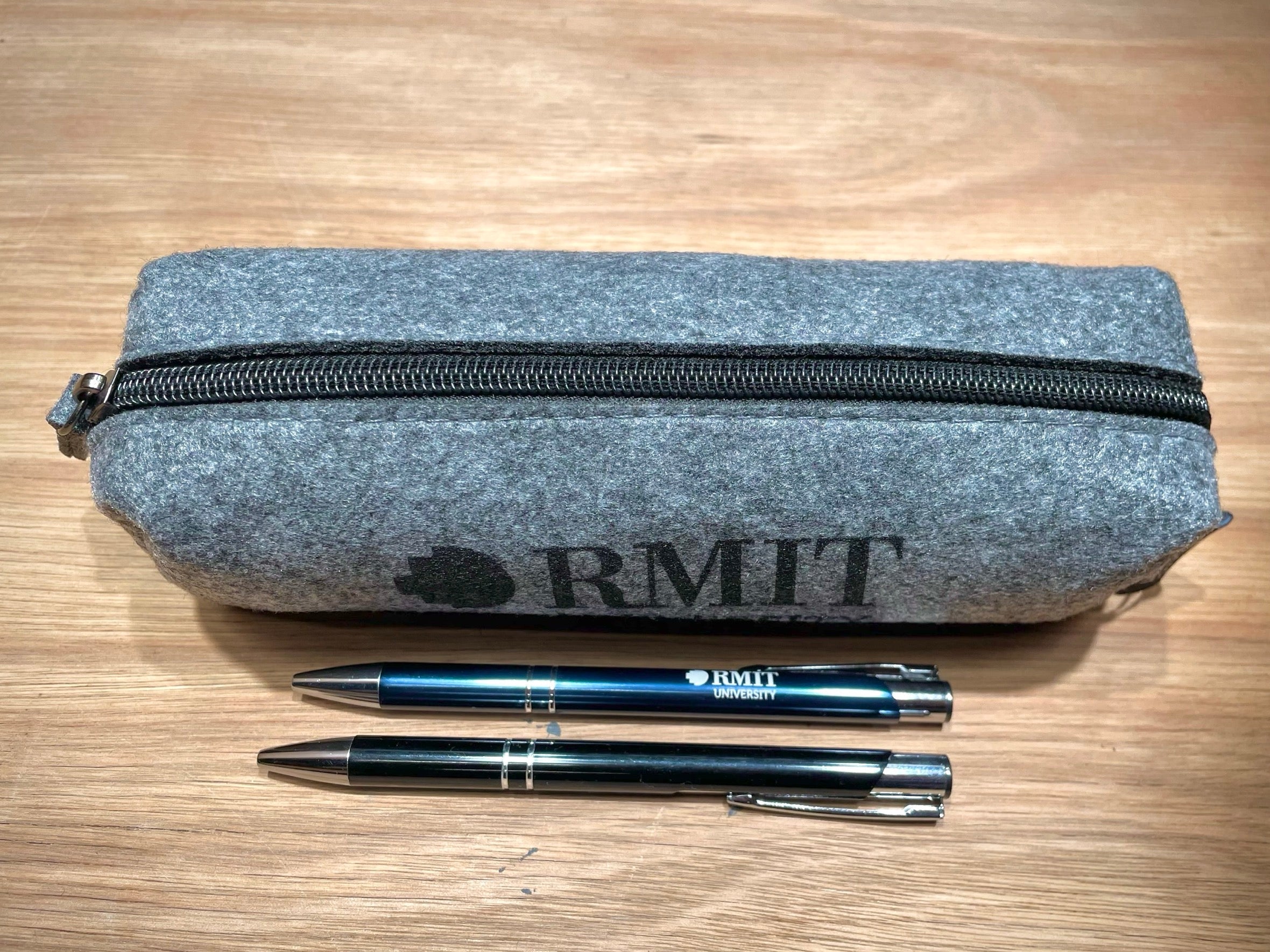 Top view of the RMIT Store rPET Utility Case in charcoal, featuring black RMIT branding printed on the front. Two pens, one black and one navy blue, are positioned in front of the case on a plain surface. The case shows its smooth textured fabric and closed zipper.