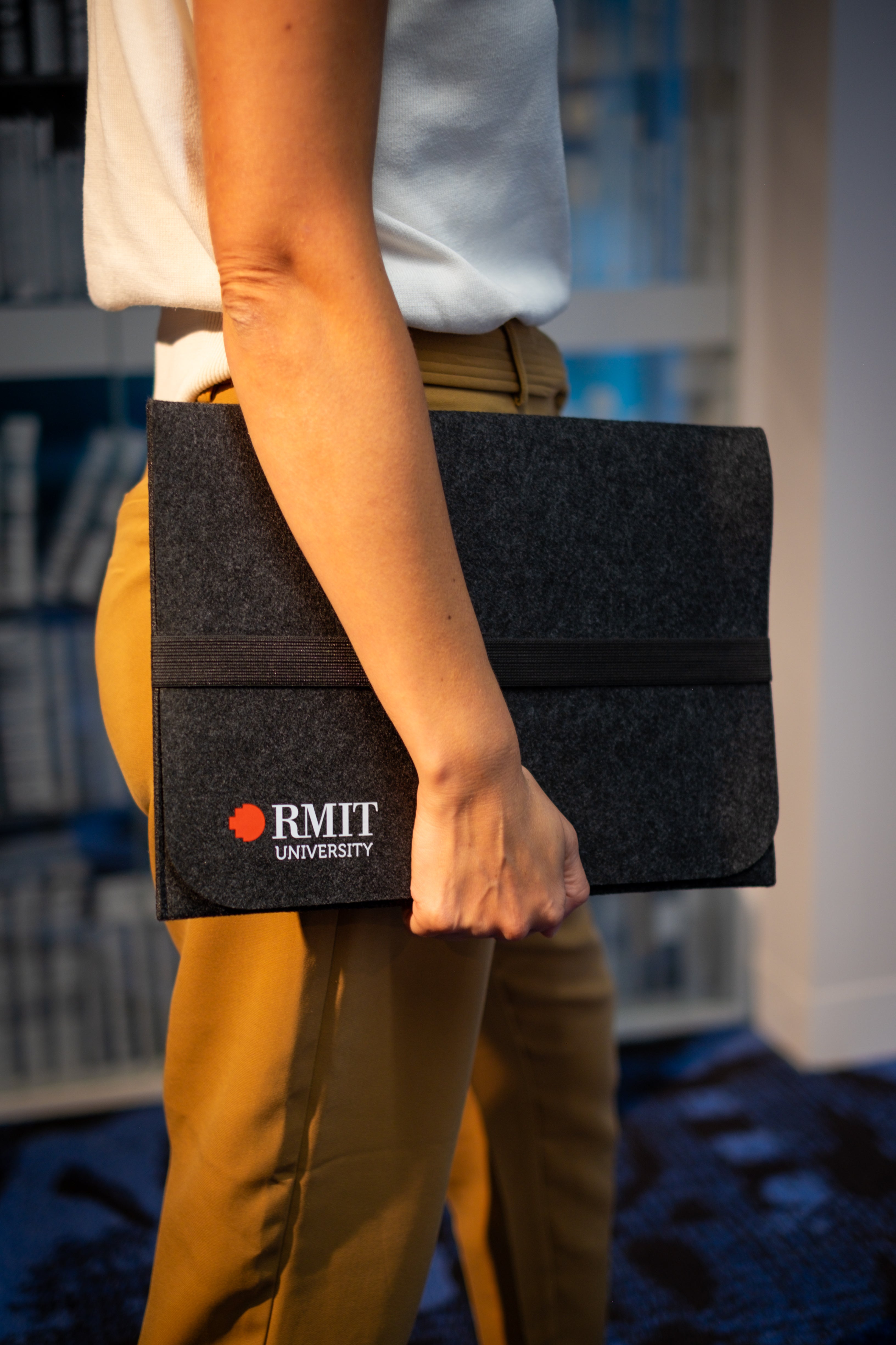 Model holding a dark grey rPET Utility Laptop Sleeve with RMIT University branding from the RMIT Store. The sleeve features a sleek design with a black elastic band, designed for eco-conscious consumers, complementing the casual brown trousers worn by the model.
