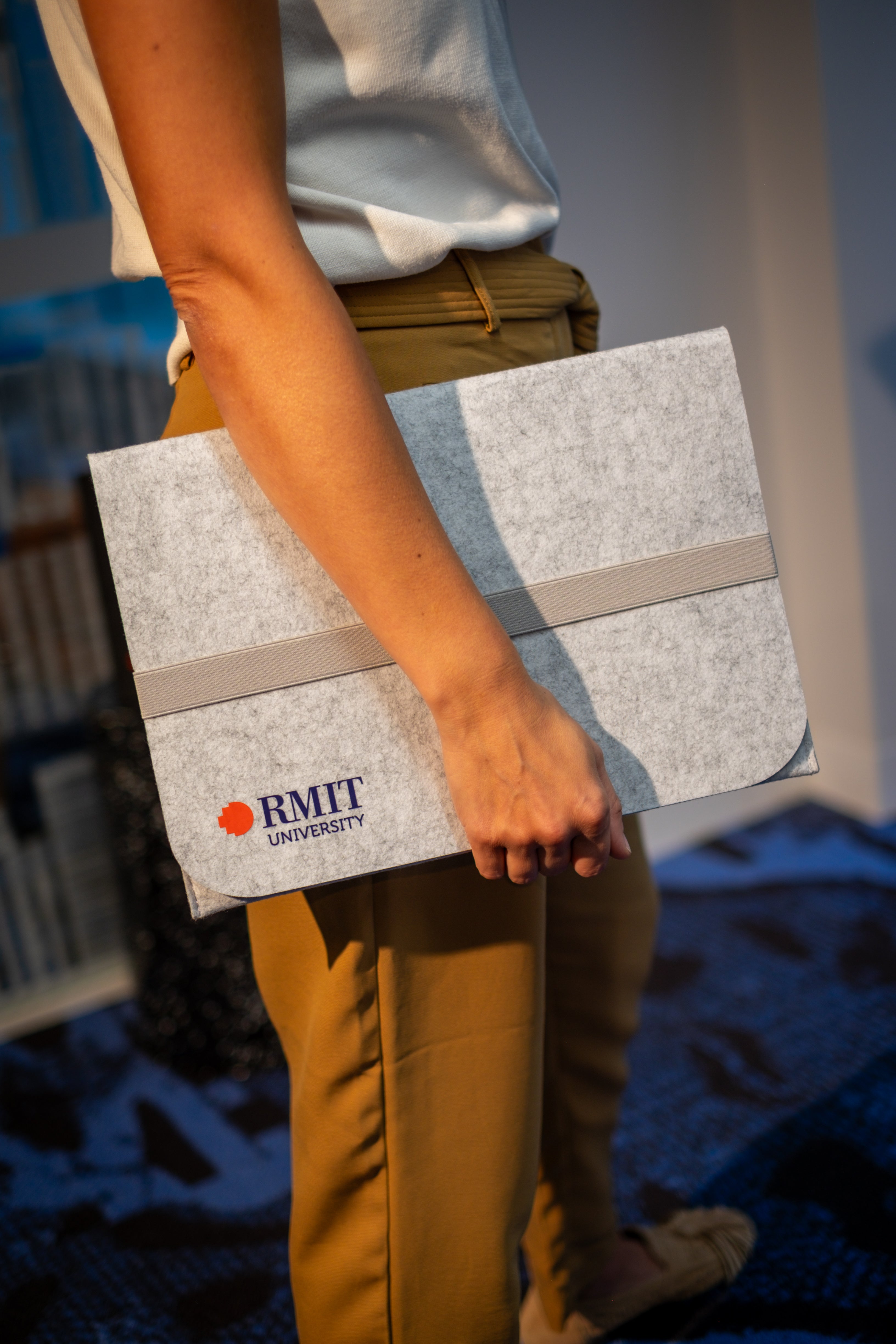 Model holding a light grey rPET Utility Laptop Sleeve with RMIT University branding from the RMIT Store. This eco-friendly sleeve has a light grey elastic band, designed for modern style, complementing the casual brown trousers worn by the model.