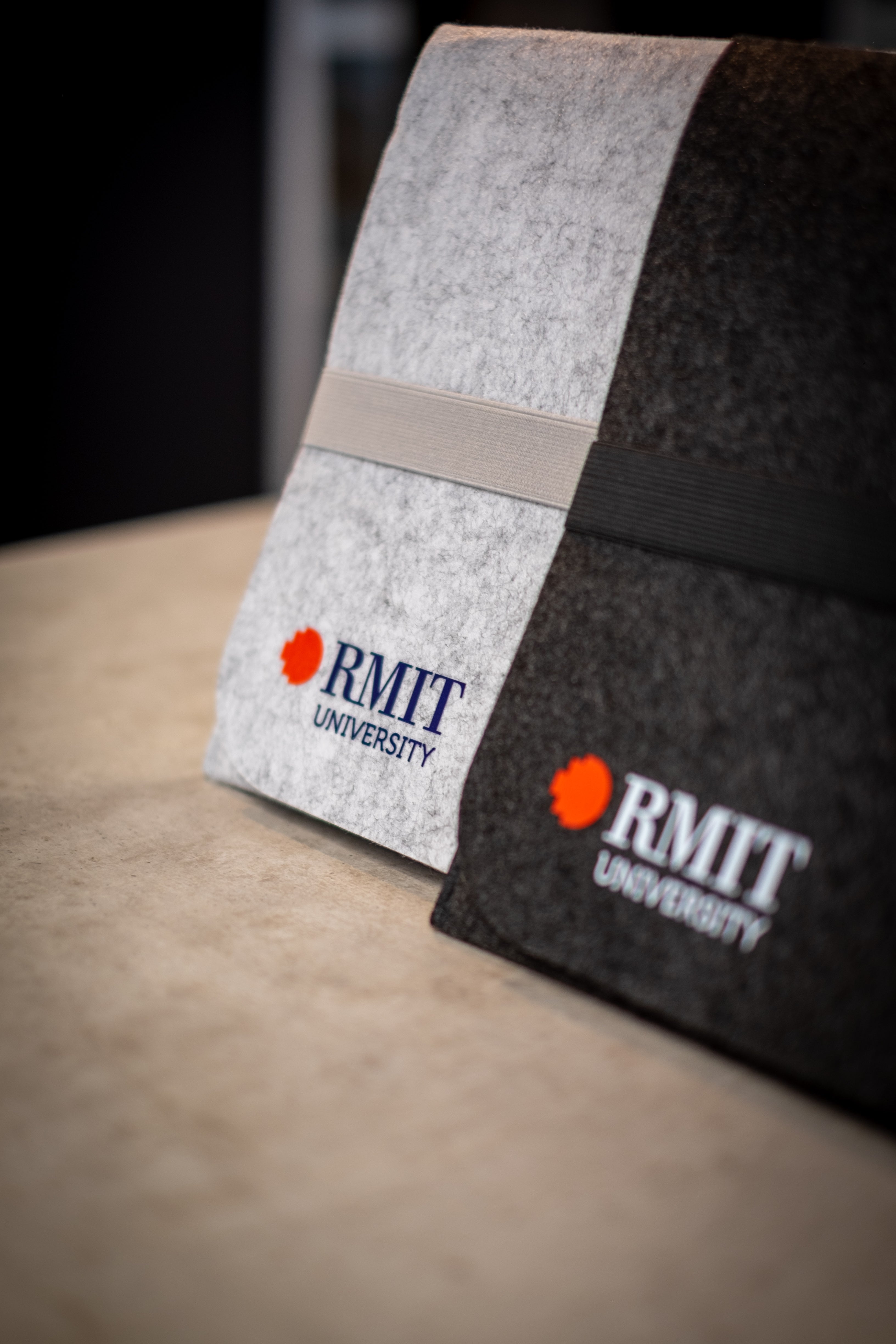 Image of two sustainable rPET laptop sleeves from RMIT Store, made from recycled plastic felt. The light grey sleeve features RMIT University's branding in red and blue, while the dark grey sleeve showcases the branding in red and white. Both sleeves have elastic bands and are designed for eco-friendly protection of devices.