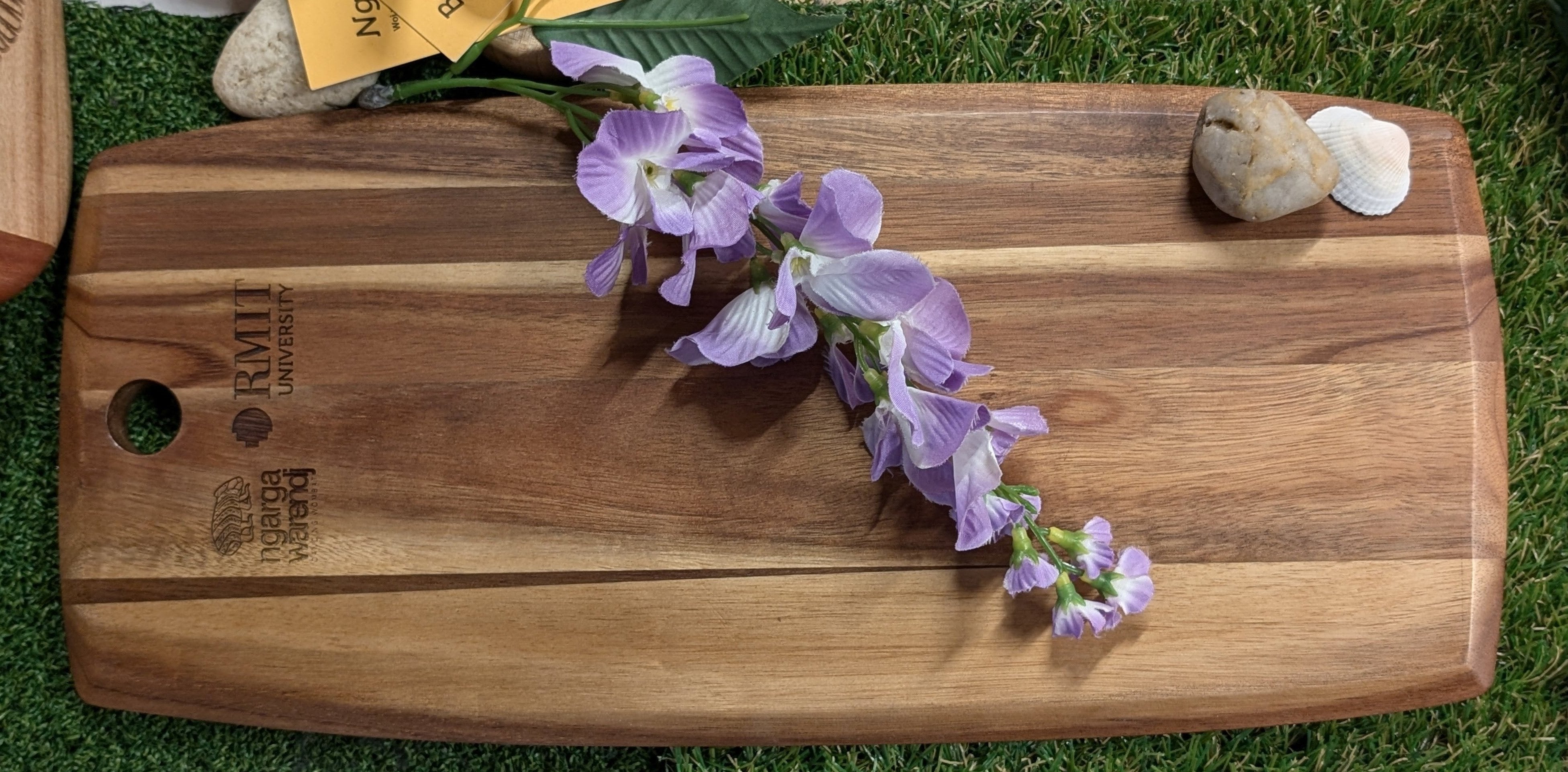 Ngarga Warendj Indigenous Australian designed Kookaburra engraved Acacia Wood Platter.  Product backing shown with RMIT University branding, lying flat with flower, rock and shell. 