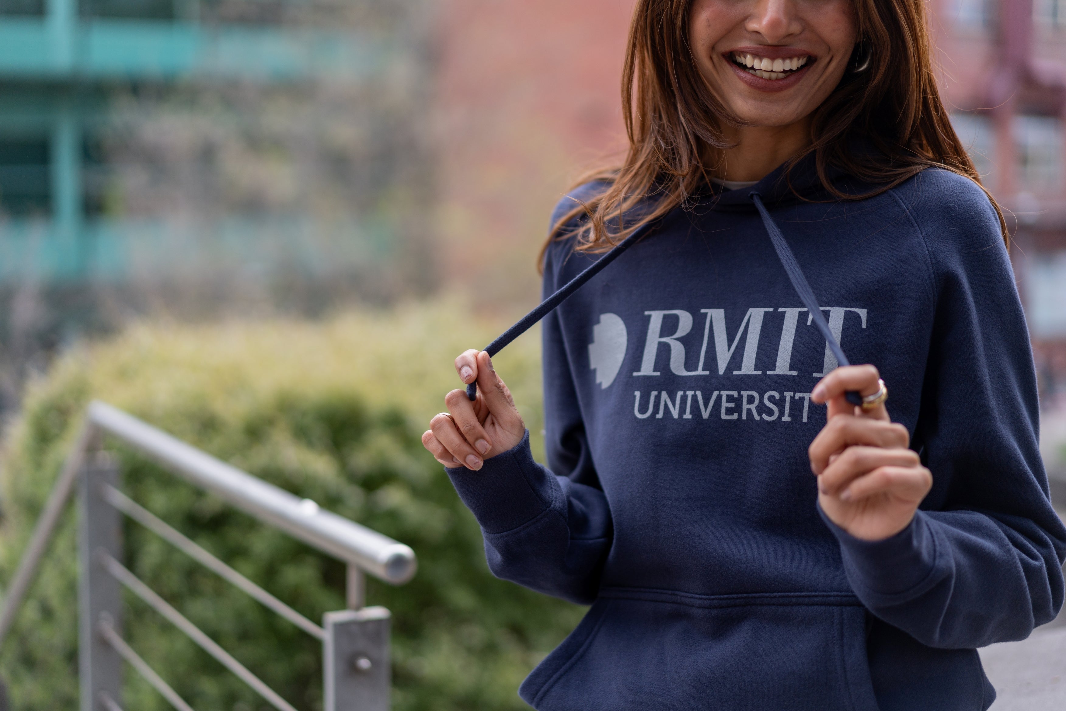 The RMIT Store's Blue Signature Hoodie featuring a grey screen printed RMIT logo. Modelled by female standing in RMIT Grounds photographed up close on the logo.
