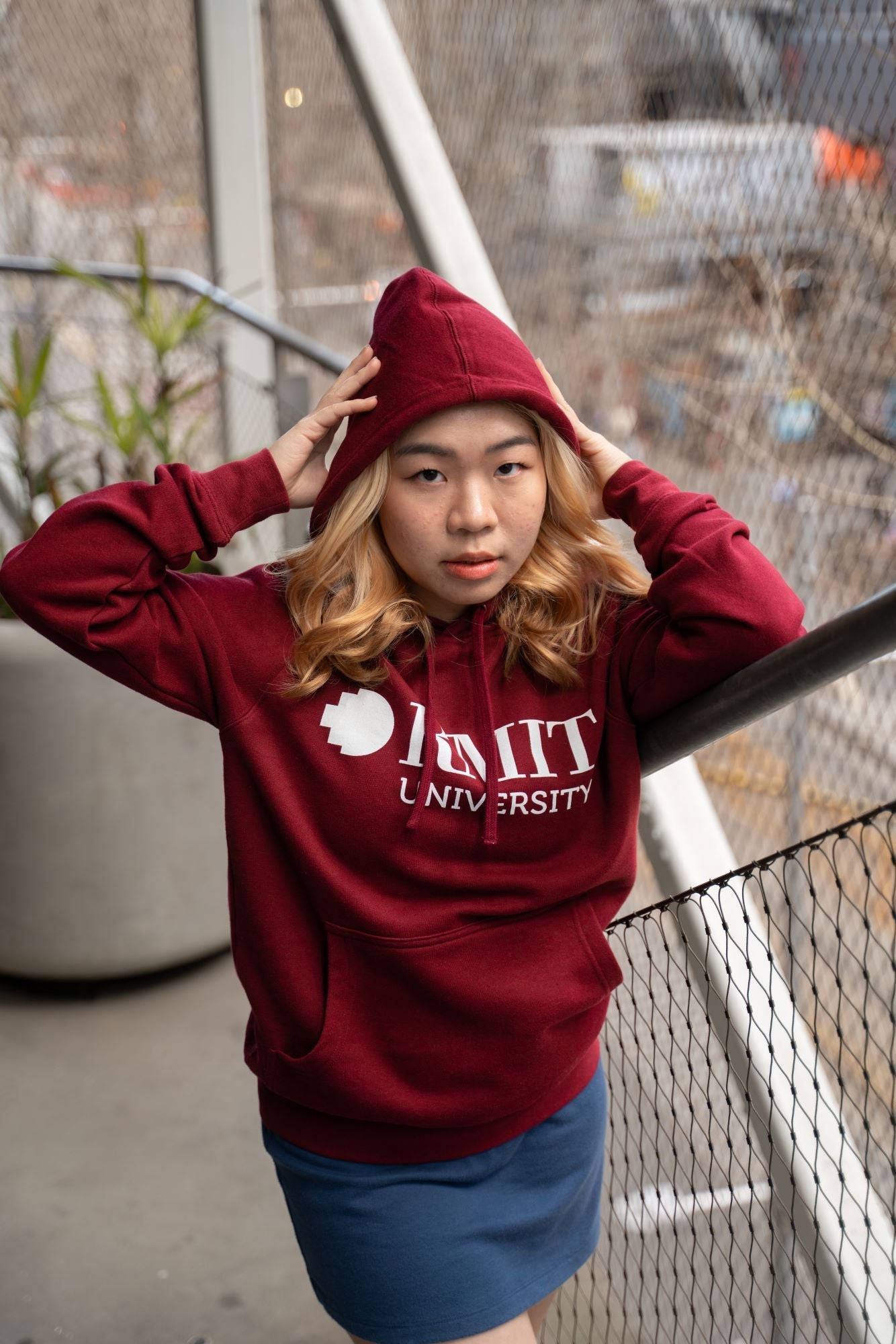 The RMIT Store's Burgundy Signature Hoodie featuring a white screen printed RMIT logo. Modelled by female standing in RMIT Grounds with the hood up photographed from the front.
