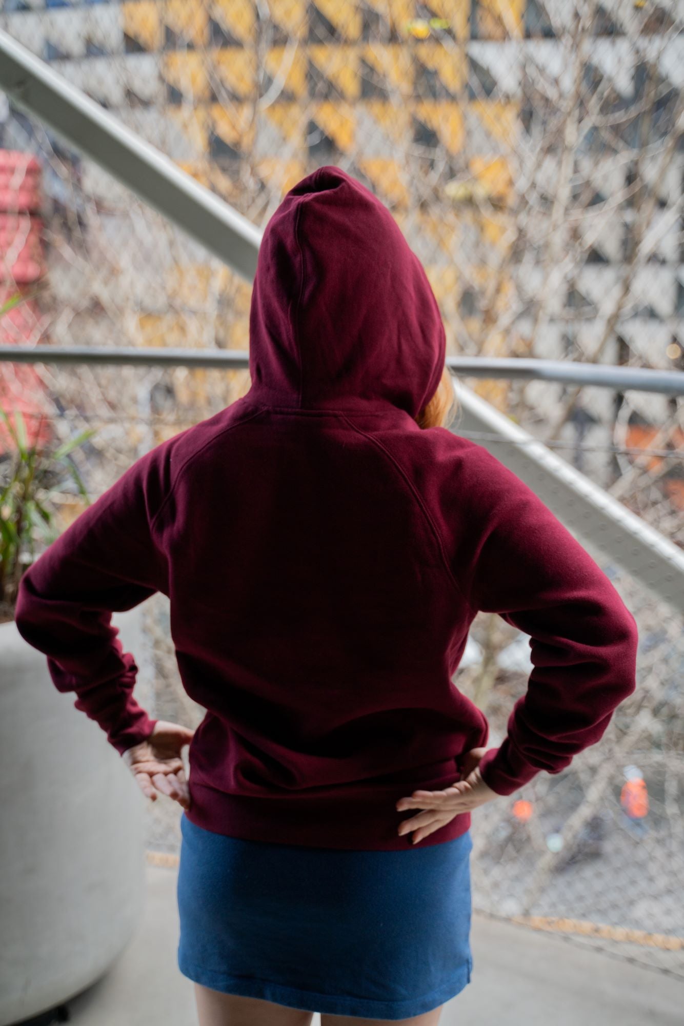 The RMIT Store's Burgundy Signature Hoodie featuring a white screen printed RMIT logo. Modelled by female standing in RMIT Grounds with the hood up photographed from the rear.
