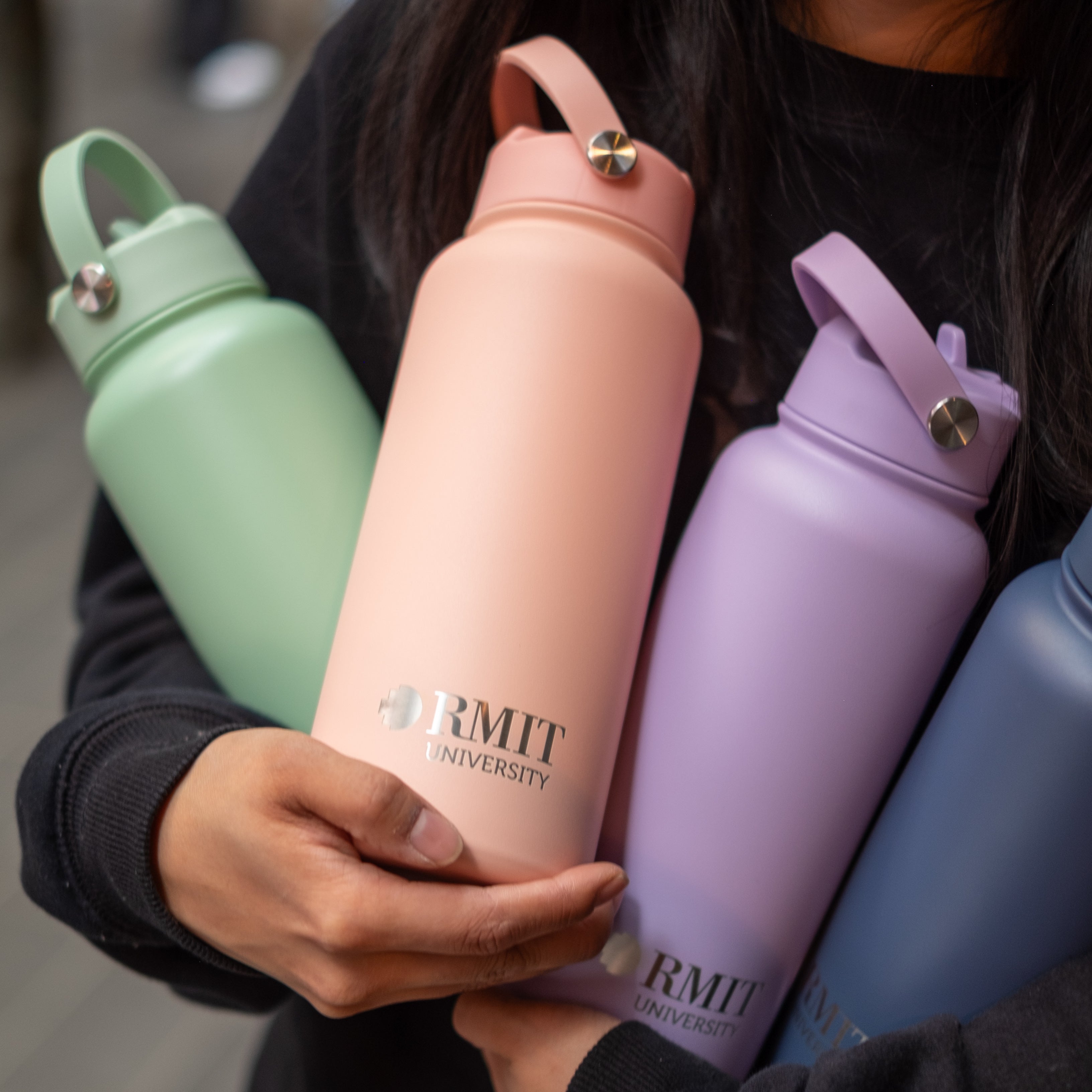 Person holding multiple pastel-coloured RMIT University stainless steel water bottles in Sage, Peach, Lilac, and Petrol Blue. Each bottle features a matte finish, a screw-top lid with a carry handle, and the RMIT University logo in silver.