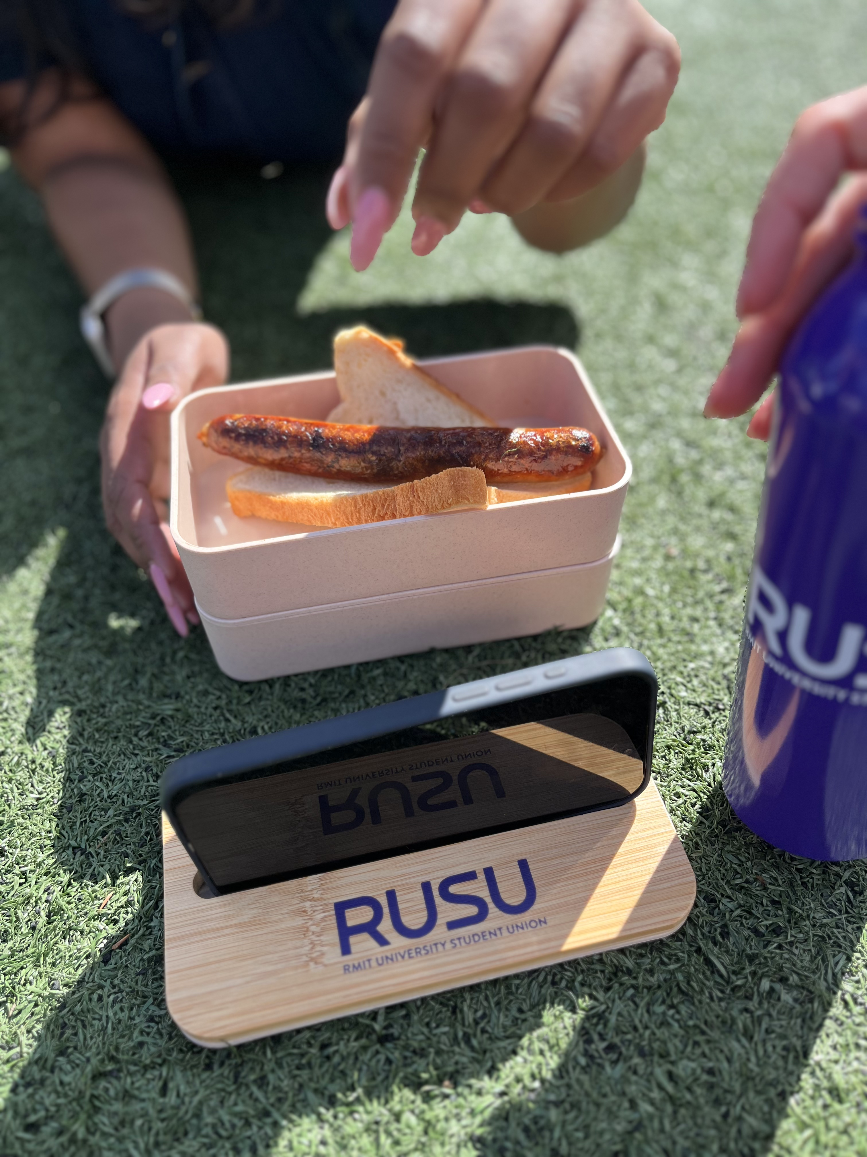 RUSU Lunchbox with two tier storage and logo printed lid with smartphone holder. Modelled in use by student with food and watching their phone at lunch time.