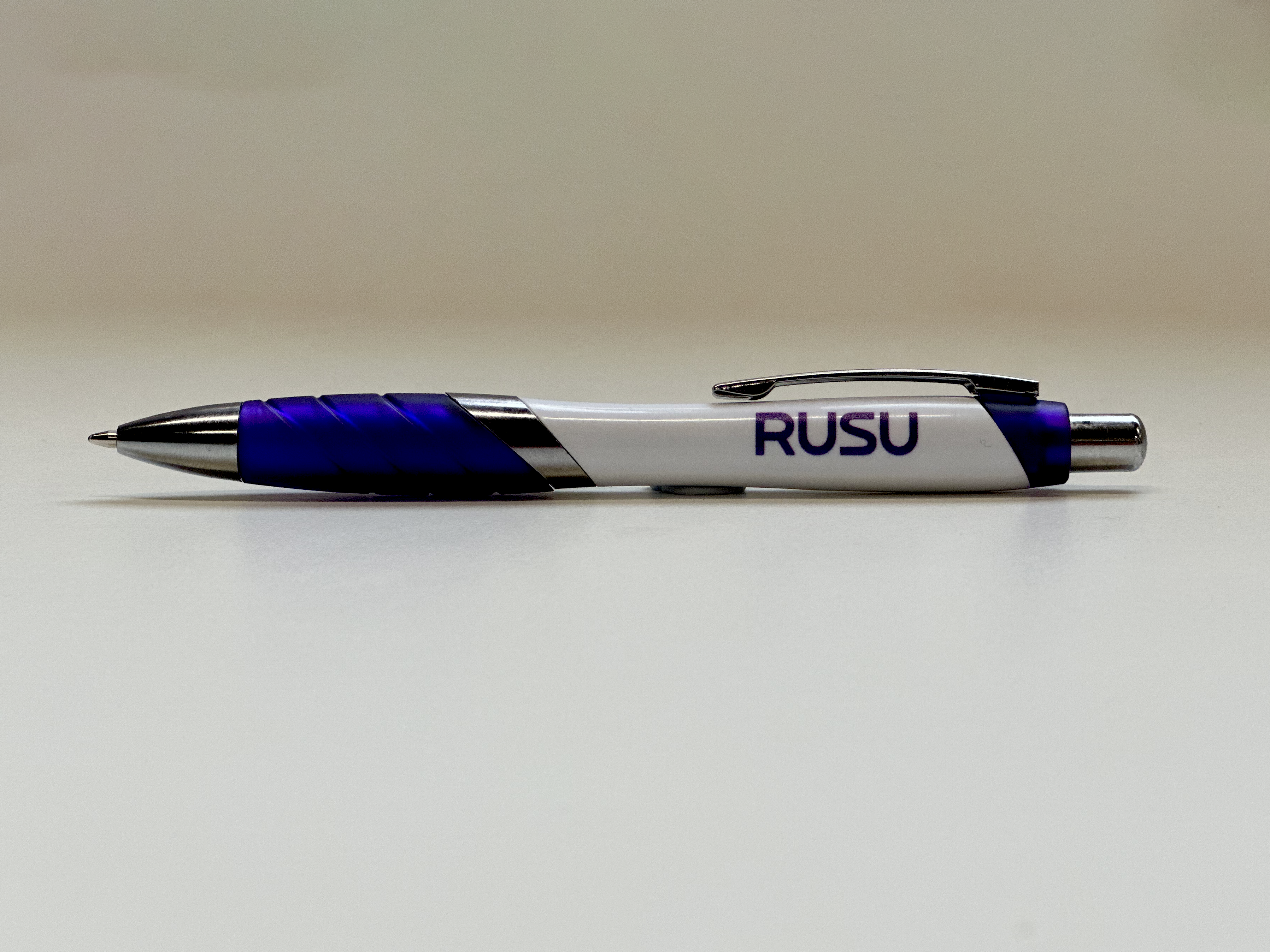 RUSU Pen in white and purple with a purple RUSU logo. Side view lying flat on surface.