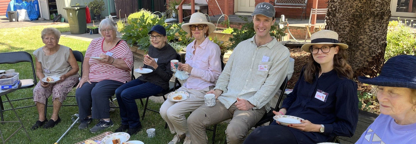 Group of St Josephs Outreach volunteers sitting outdoors on chairs, enjoying cake with plates and cups.