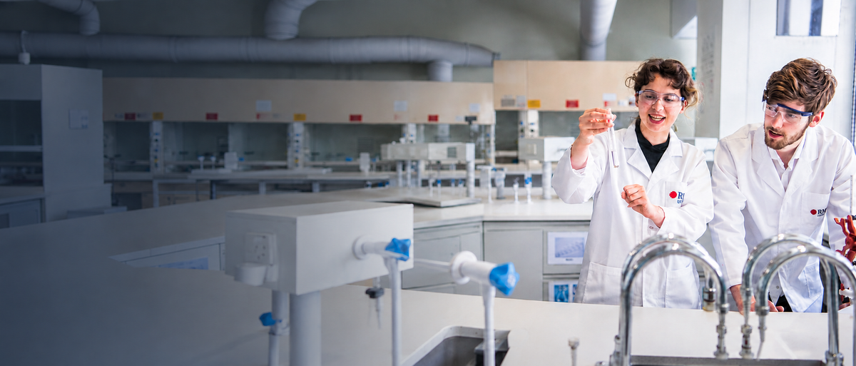 RMIT Store Lab Coats in white and clear Safety Glasses, modelled from the front by two models at a science laboratory bench, with each long-sleeved coat featuring a buttoned front, large pockets and a coloured RMIT University logo on the left chest; one model holds a glass test tube while the other watches.