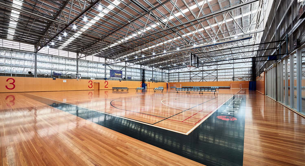 An indoor basketball court with polished wooden floors, orange walls, and a high industrial metal roof with skylights. At RMIT Sport Centre Bundoora