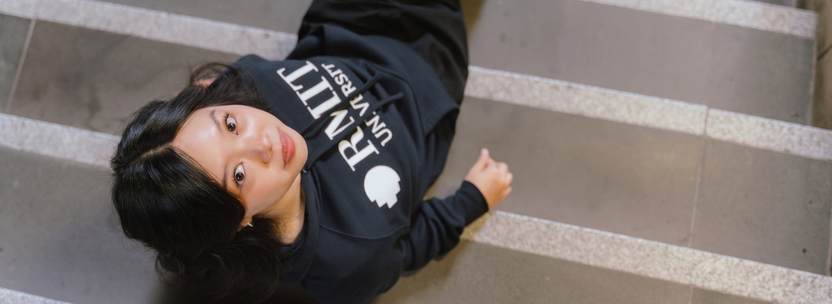 Top-down view of model lying on indoor staircase wearing RMIT Essential Hoodie in navy featuring bold white RMIT University logo across the chest, with hood down and relaxed fit, captured from above with strong perspective lines of steps, available at the RMIT Store.