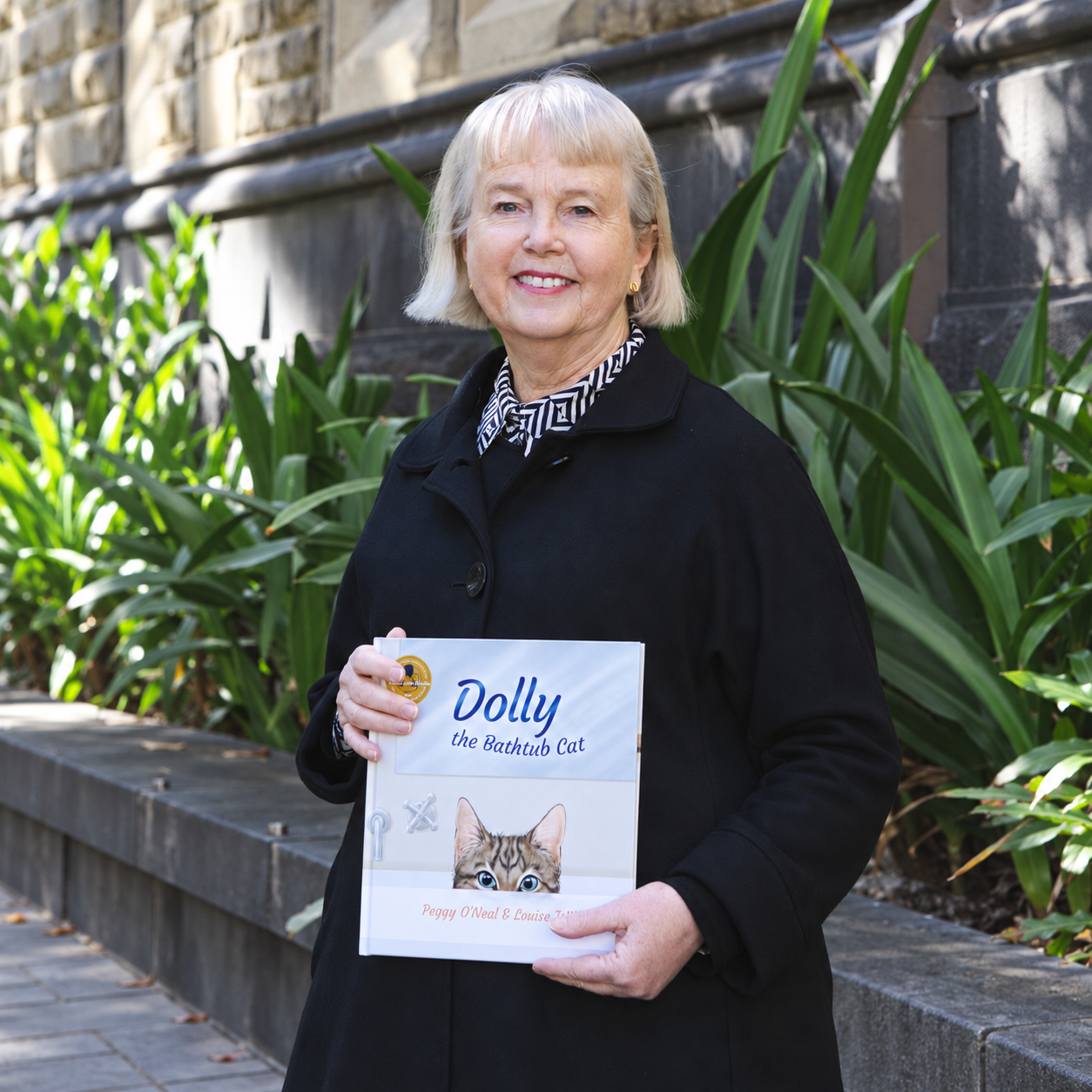 Peggy O’Neal smiling outdoors while holding a copy of the children’s book Dolly the Bathtub Cat book.
