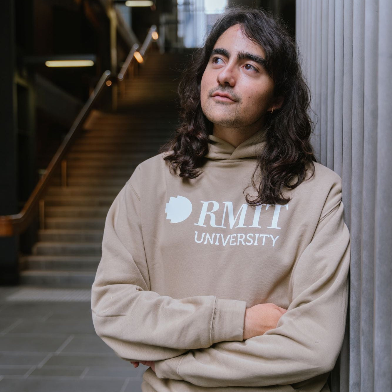 Student leaning against a concrete pillar wearing RMIT Casey Hoodie in sand with large white RMIT University logo across the chest, hood down with long hair visible, arms crossed, styled with dark pants in an indoor campus stairwell setting.